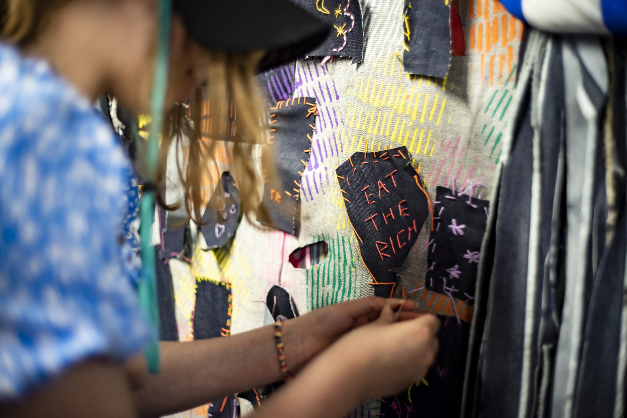 A person stitching on a multicoloured fabric wall, adding a patch with orange thread that reads EAT THE RICH in capital letters. The person's face is not shown clearly.