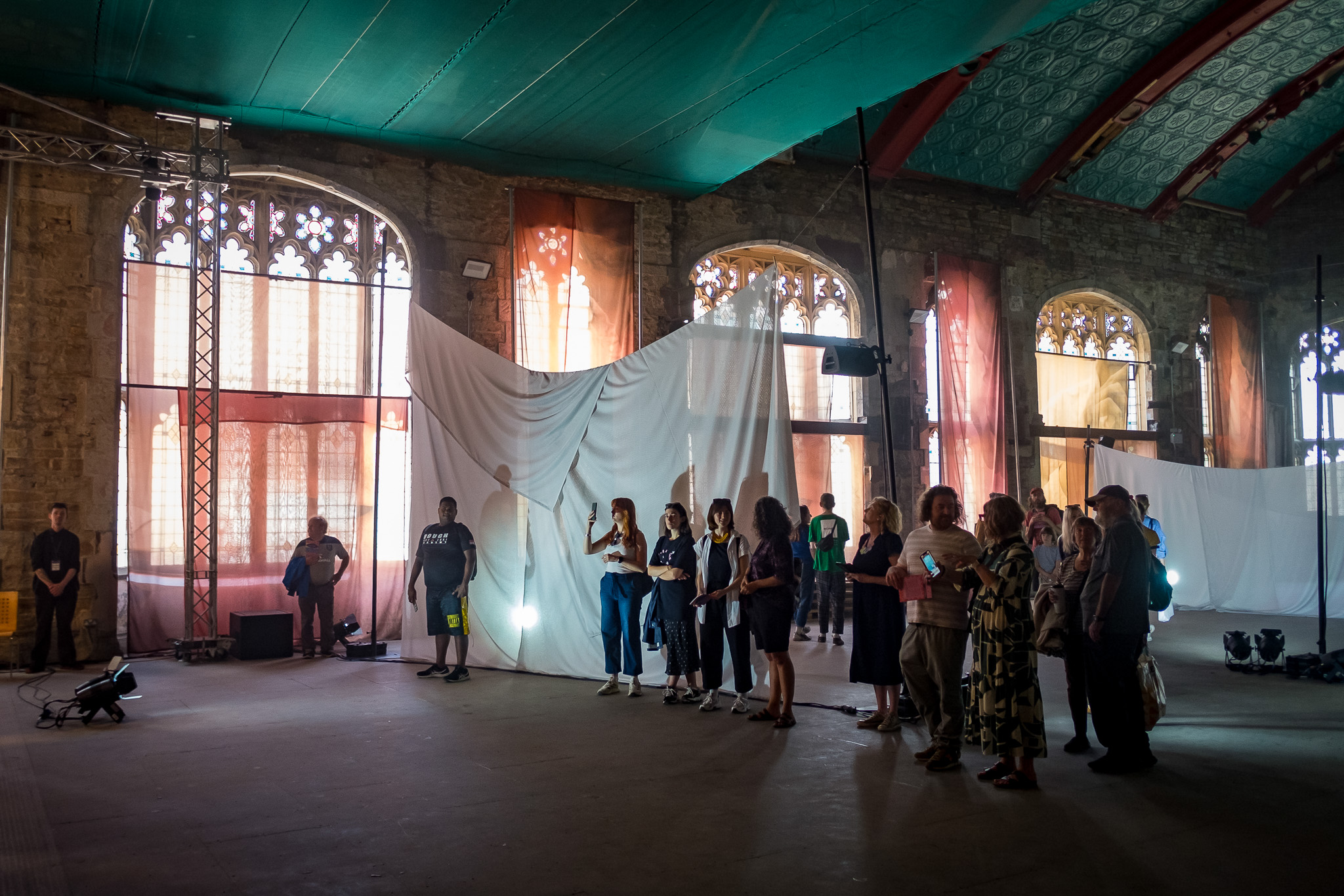 A group of people stands together indoors in a large, high-ceilinged room with arched windows, draped fabric, and coloured lighting, suggesting an art exhibition or event.