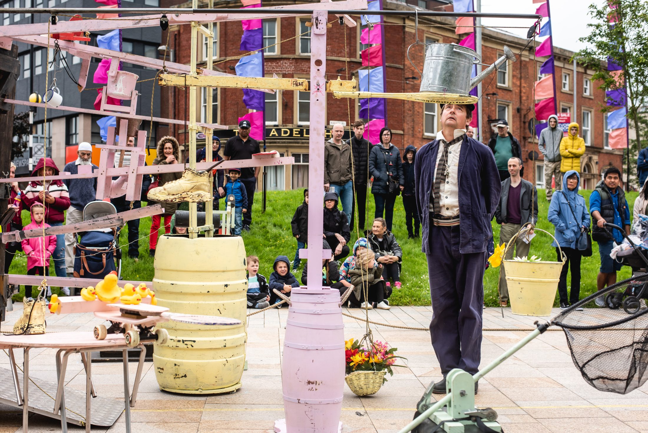 Performer balancing a metal bucket on his head amid a colorful contraption, while onlookers, including children, watch curiously.