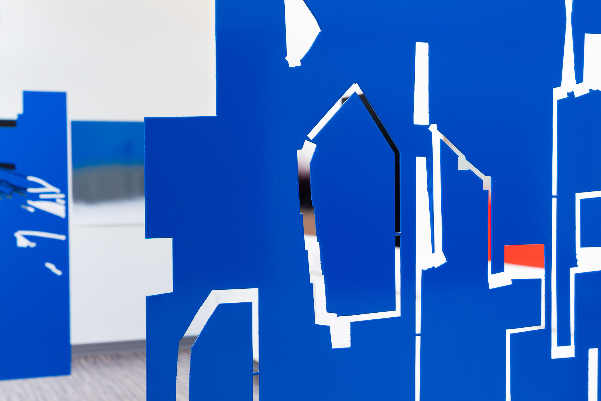 Bright blue abstract sculptures with irregular geometric cut-outs are displayed in a modern, white-walled gallery space. Blurred art pieces hang in the background, and some orange accents are visible through the cut-outs.