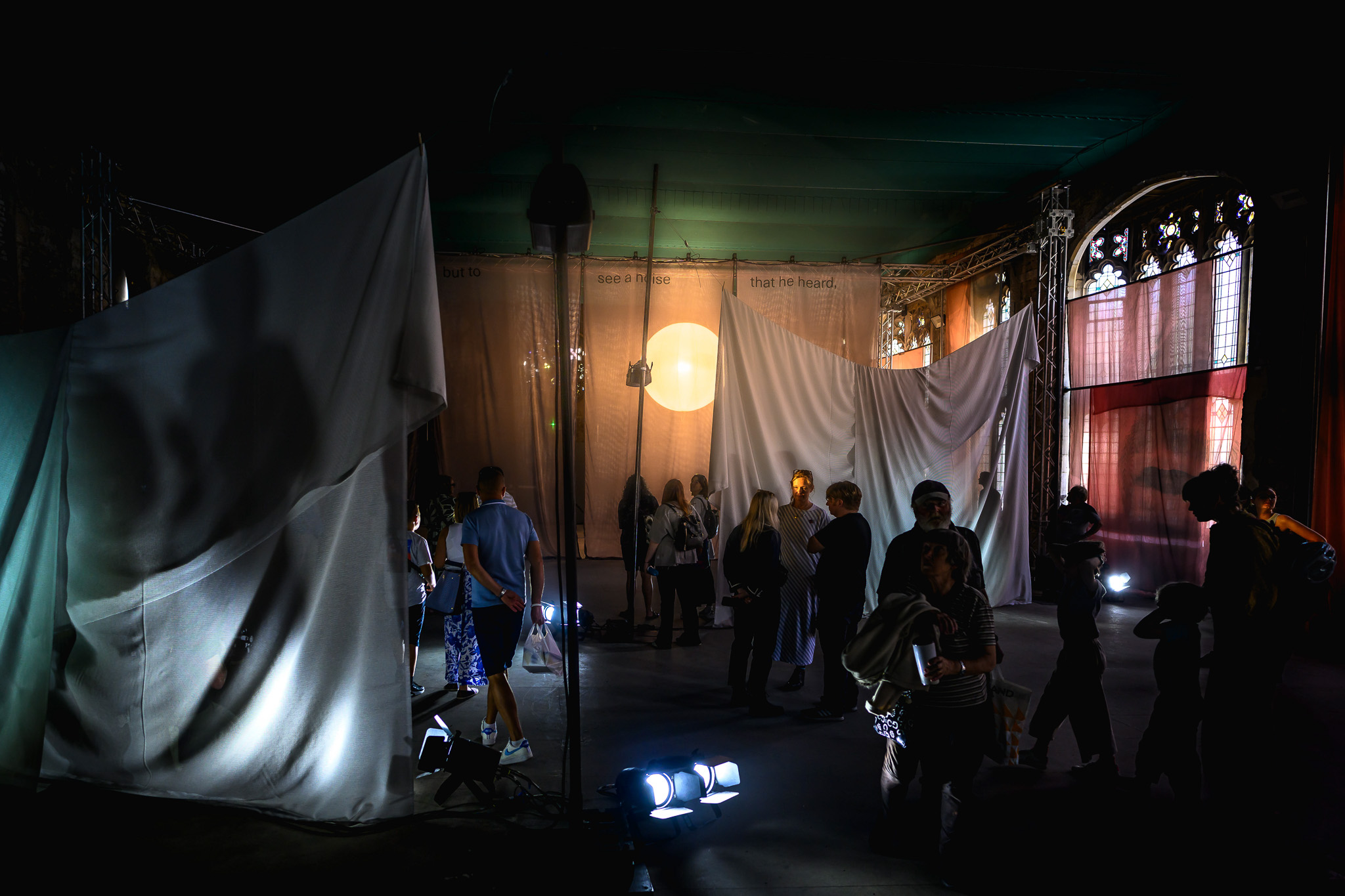 A group of people stand in a dimly lit room with draped white sheets, spotlights on the floor, and sunlight streaming through windows, creating dramatic shadows and a mysterious atmosphere.