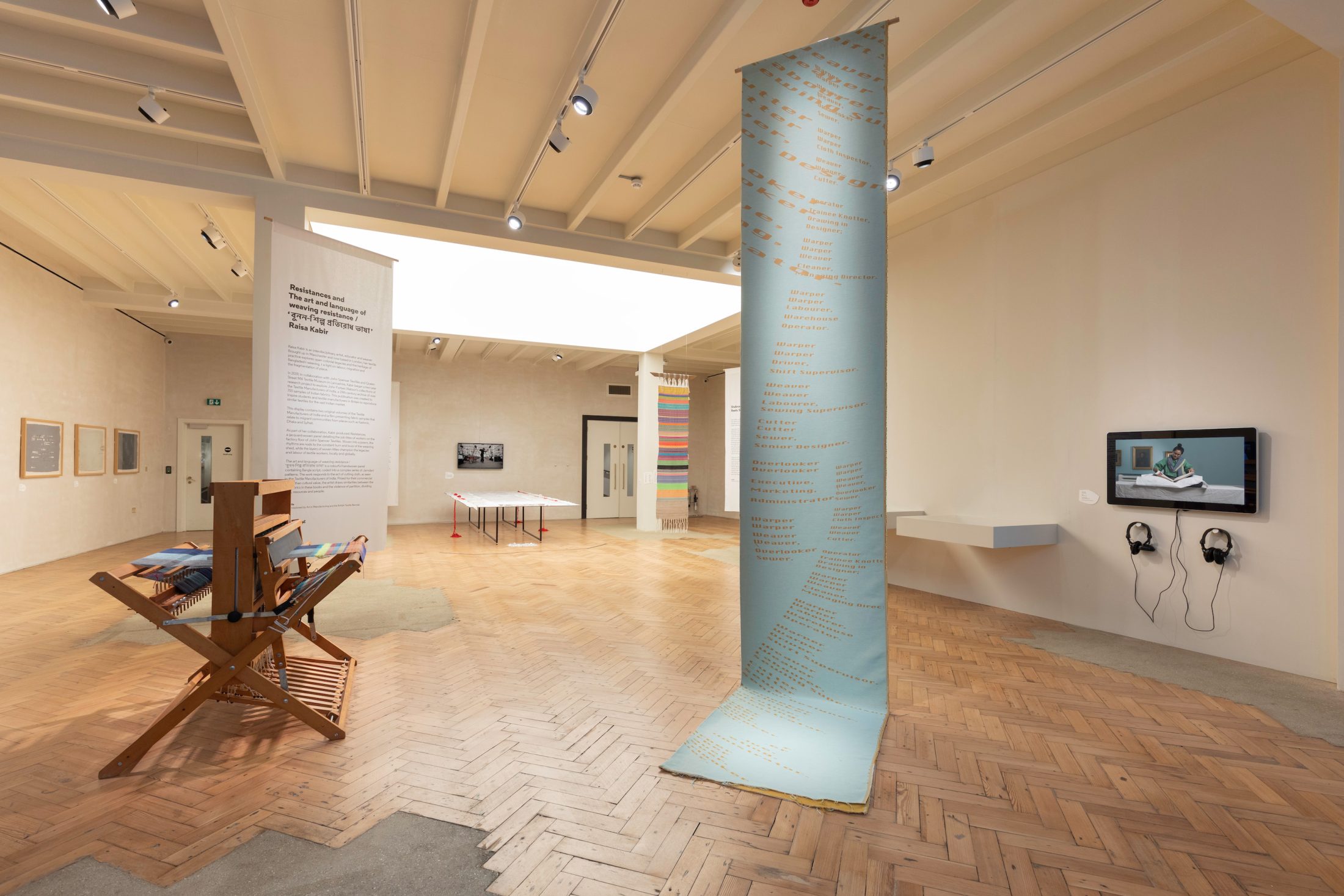 A contemporary art gallery with wooden floors displays installations, textiles, framed artworks, and a tall blue vertical banner with text. There’s a video screen with headphones on the wall and soft natural lighting from above.
