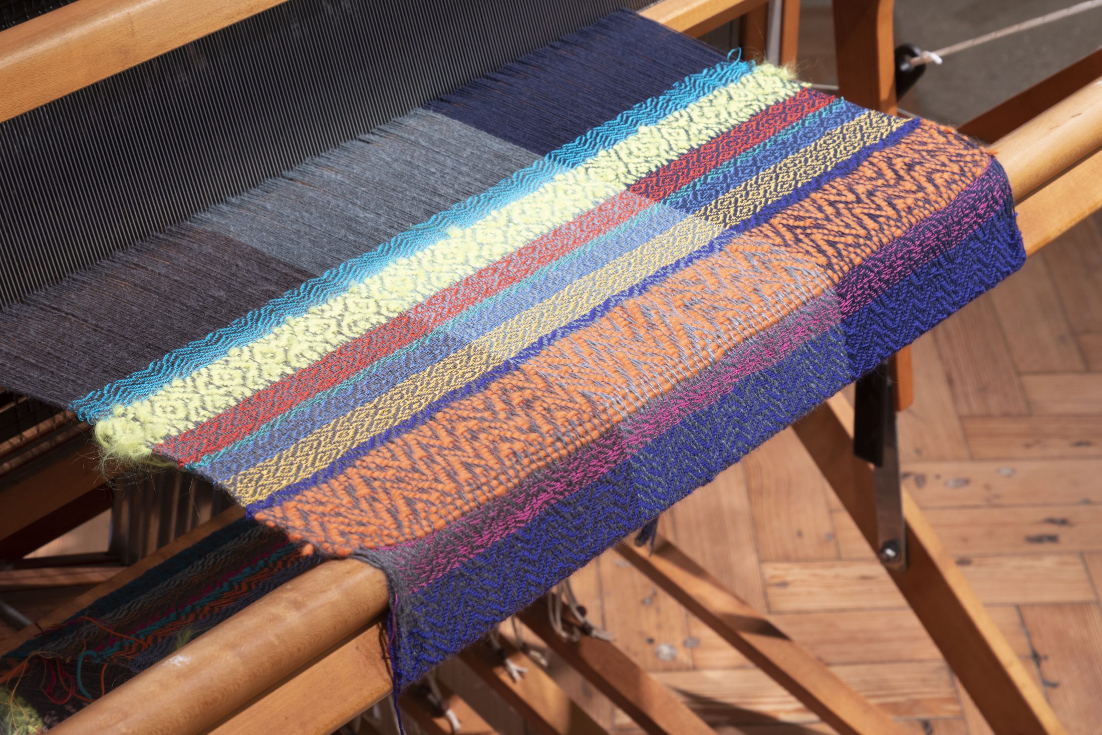 A close-up of a colourful woven textile on a wooden loom, featuring intricate patterns in blue, orange, yellow, and green threads. The loom and parquet flooring are partially visible.