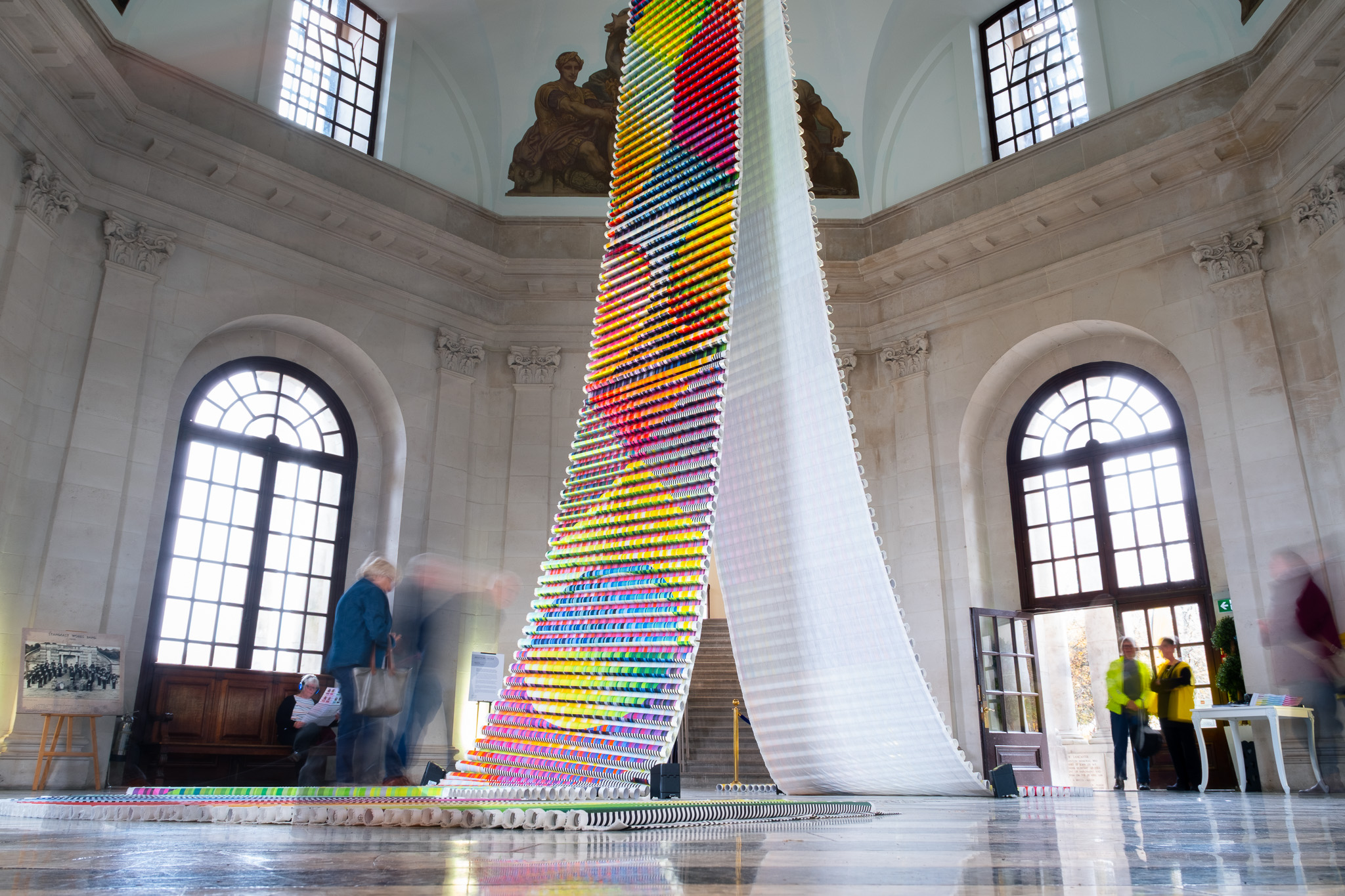 A tall, curved tower made of colourful stacked cups stands in a grand hall with large arched windows and marble floors. Several blurred people walk around, viewing the display.