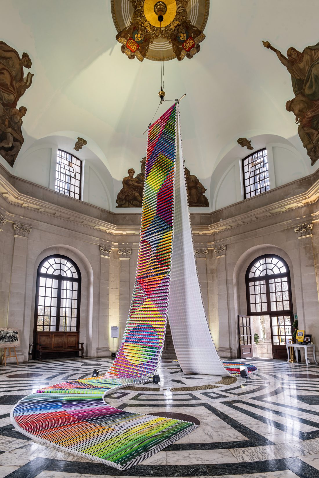 Tall, colourful woven art installation hangs from a domed ceiling in an ornate, high-ceilinged room with arched windows, marble floors, and classical paintings on the walls.