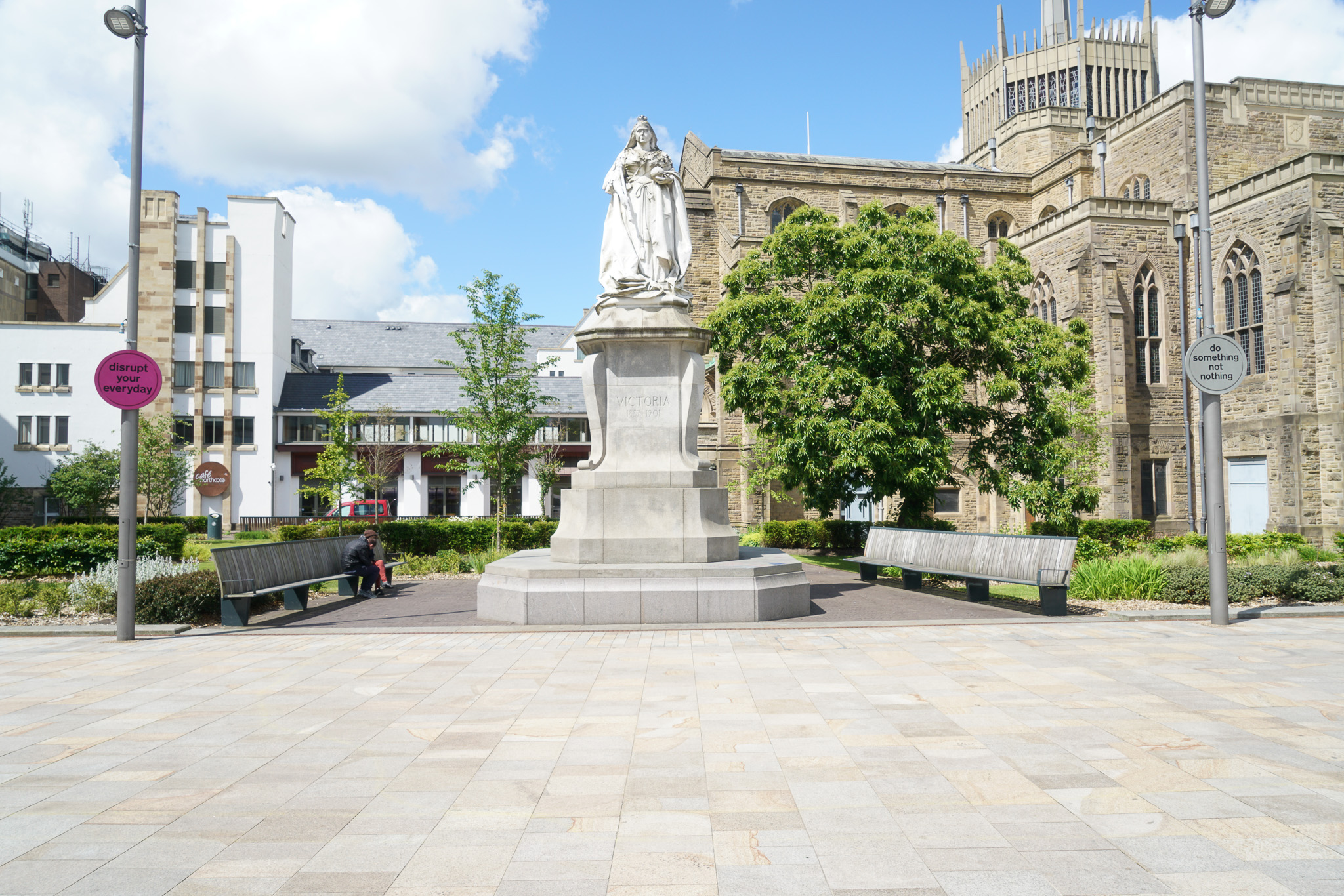 A statue stands on a stone plinth in a town square, surrounded by benches, greenery, and historic buildings under a blue sky with clouds. Signs are posted on poles beside the square.