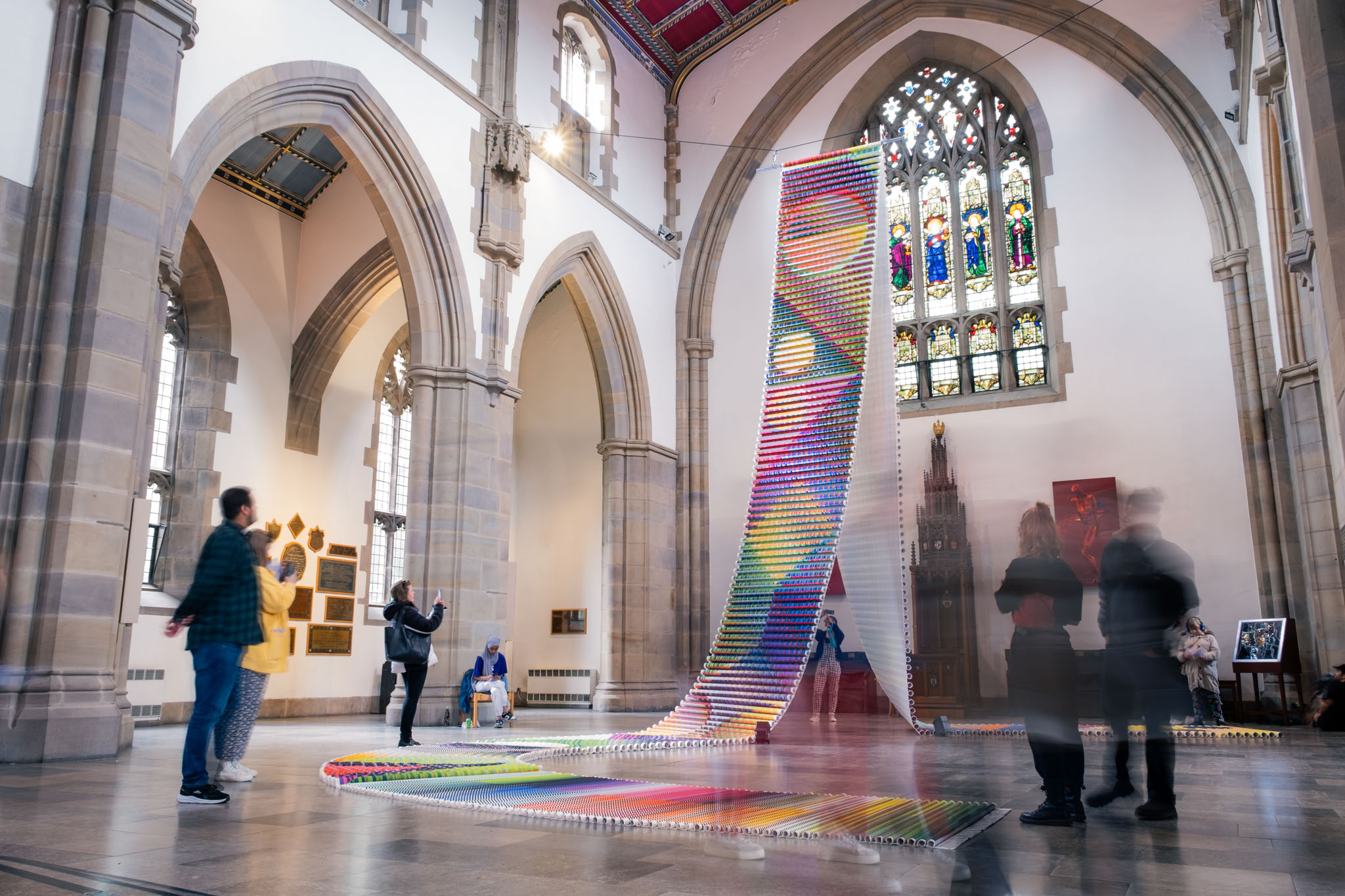 People observe a large, colourful domino art installation cascading in a wave pattern inside a grand hall with high arched windows, stained glass, and stone columns. Some visitors are blurred, suggesting movement.