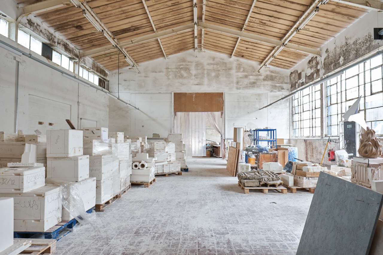 A spacious, high-ceilinged workshop with large windows, filled with stacked boxes, materials, and wooden pallets. The walls and floor are dusty, suggesting ongoing building or refurbishment work.