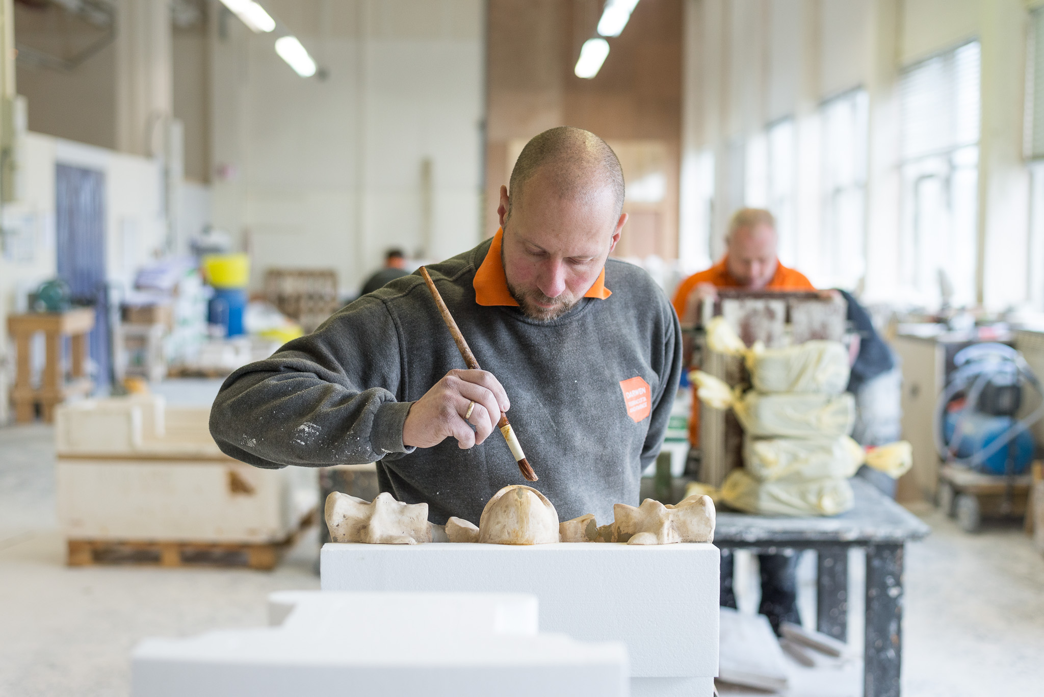 Craftsperson carefully painting a sculptural piece with a fine brush in a bright workshop, with another worker blurred in the background.