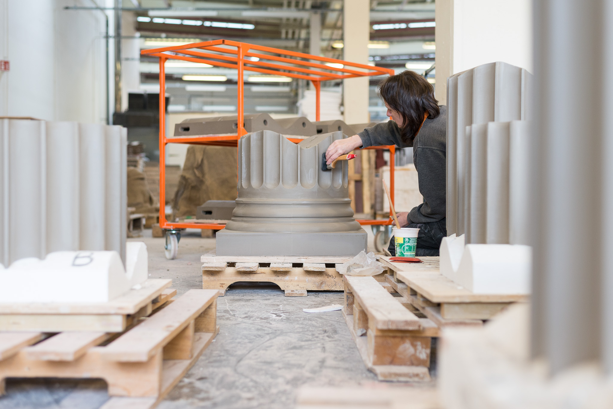 Craftsperson smoothing and finishing a large decorative column base in a workshop, surrounded by similar moulded pieces on wooden pallets.