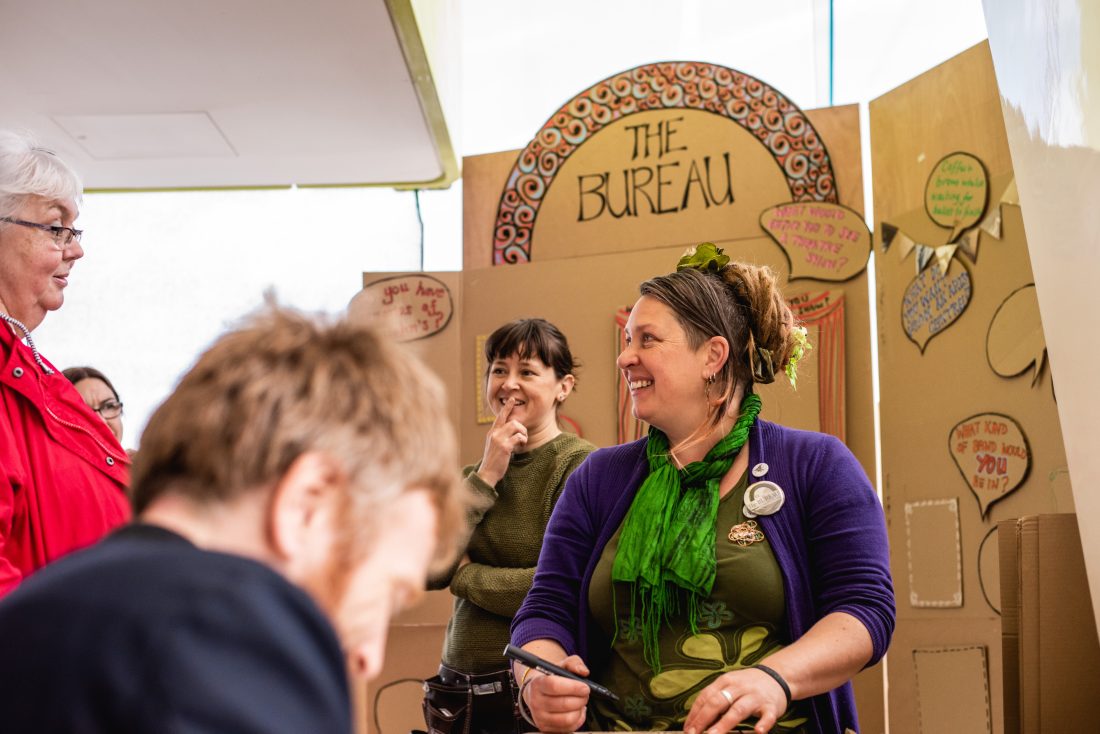 A woman in green and purple smiles and talks to people in front of a colourful cardboard stall labelled The Bureau. Others nearby are engaged and chatting in a cheerful setting.