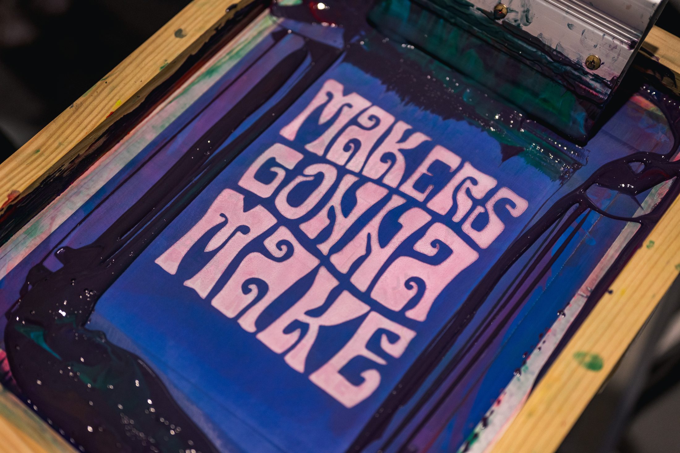 A screen printing frame with purple and blue ink displays the phrase “Makers Gonna Make” in bold, white, stylised letters on the mesh screen.