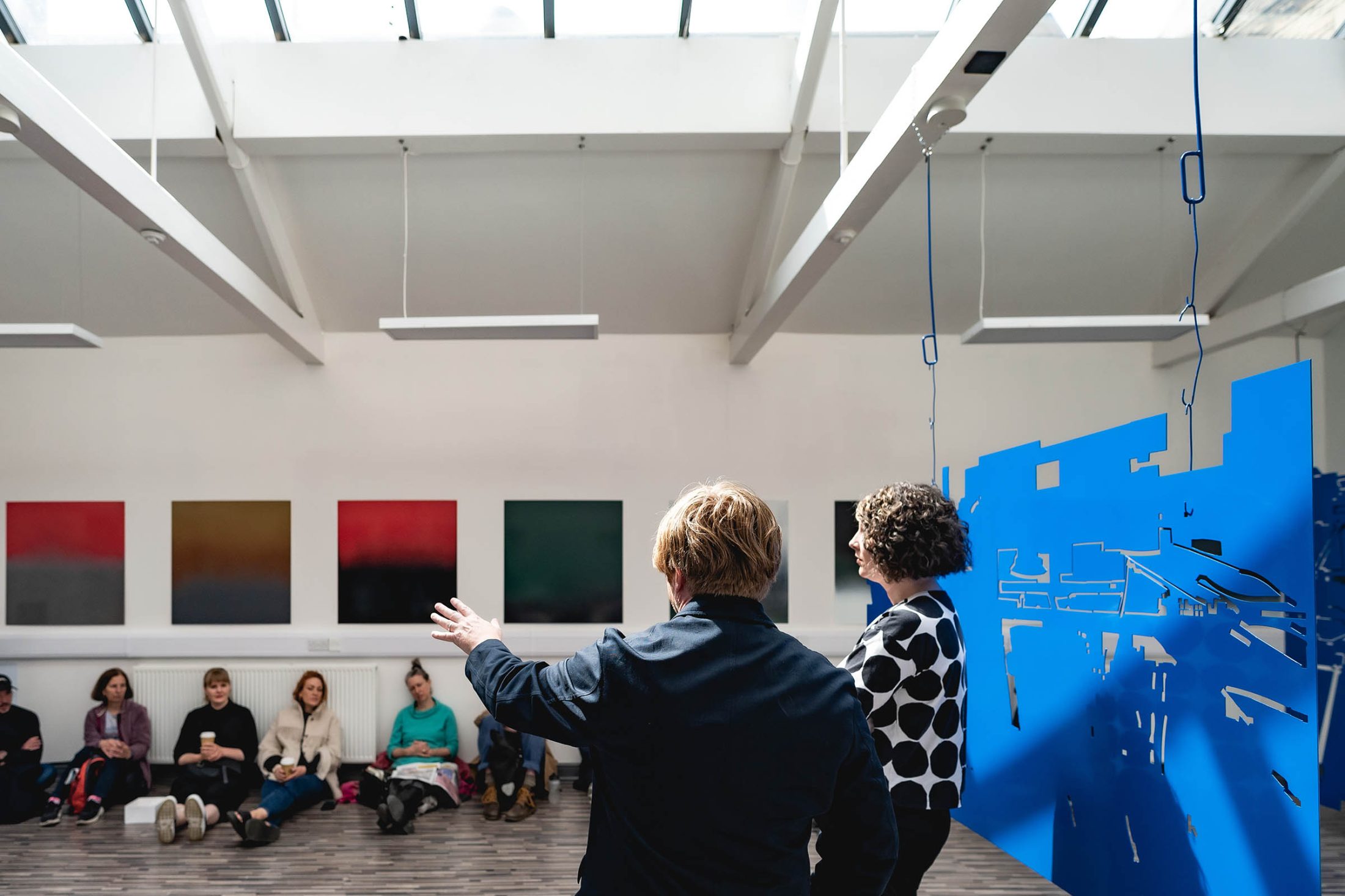 People in a modern art gallery stand and gesture toward a blue abstract installation, while seated viewers observe, with colourful paintings on the walls.