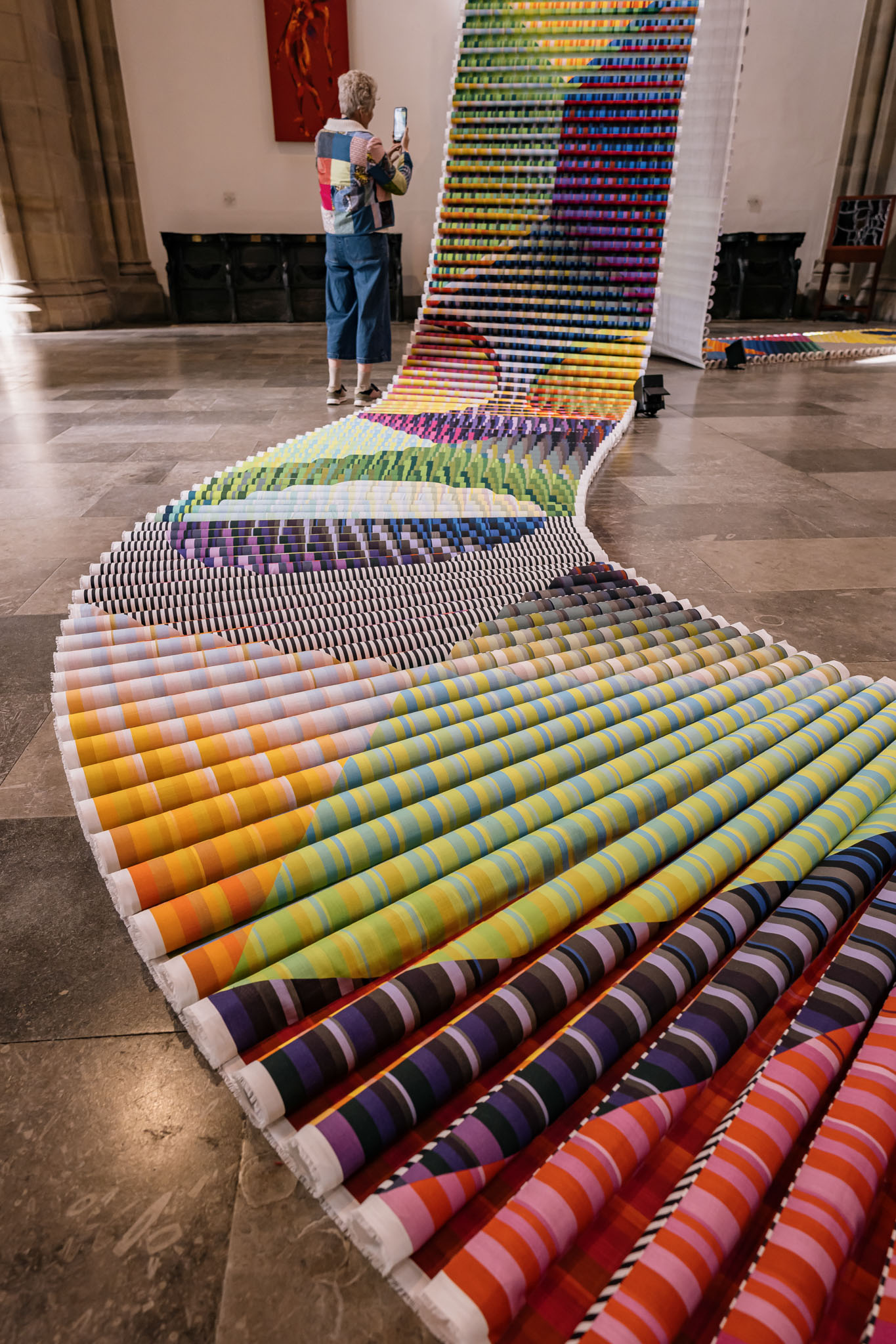 A person takes a photo of a large, colourful art installation featuring rows of rolled rainbow-striped fabric unfurling across a spacious hall with high ceilings and stone floors.
