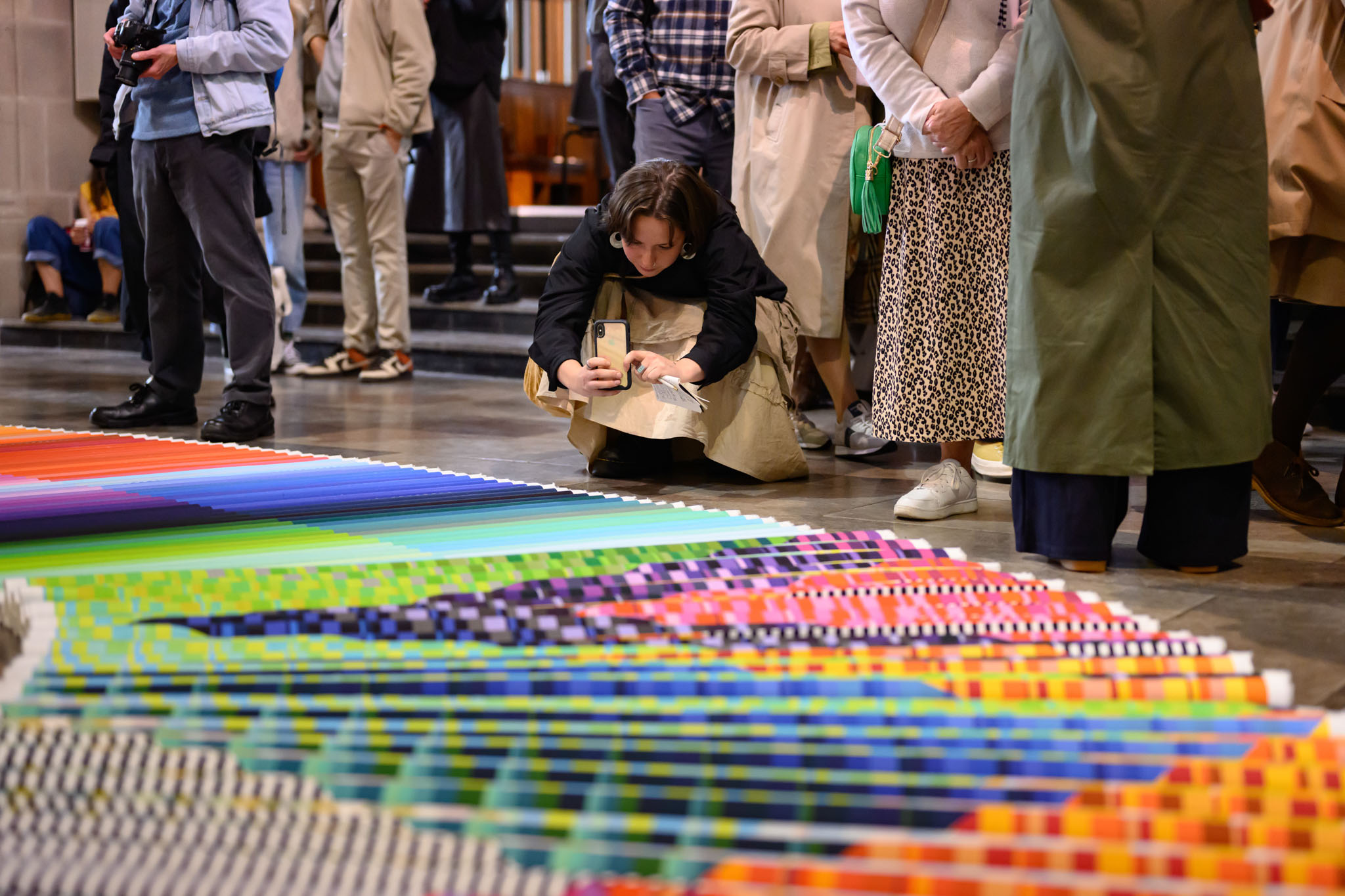 A person crouches to take a photo with their phone of a colourful, geometric artwork displayed on the floor, whilst others stand and observe around them in an indoor setting.