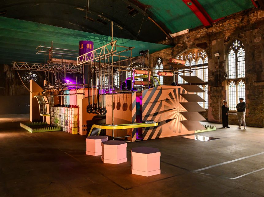 A brightly lit cardboard art installation of ropes and bars sits in a large industrial hall with arched windows and exposed brick, as visitors observe.
