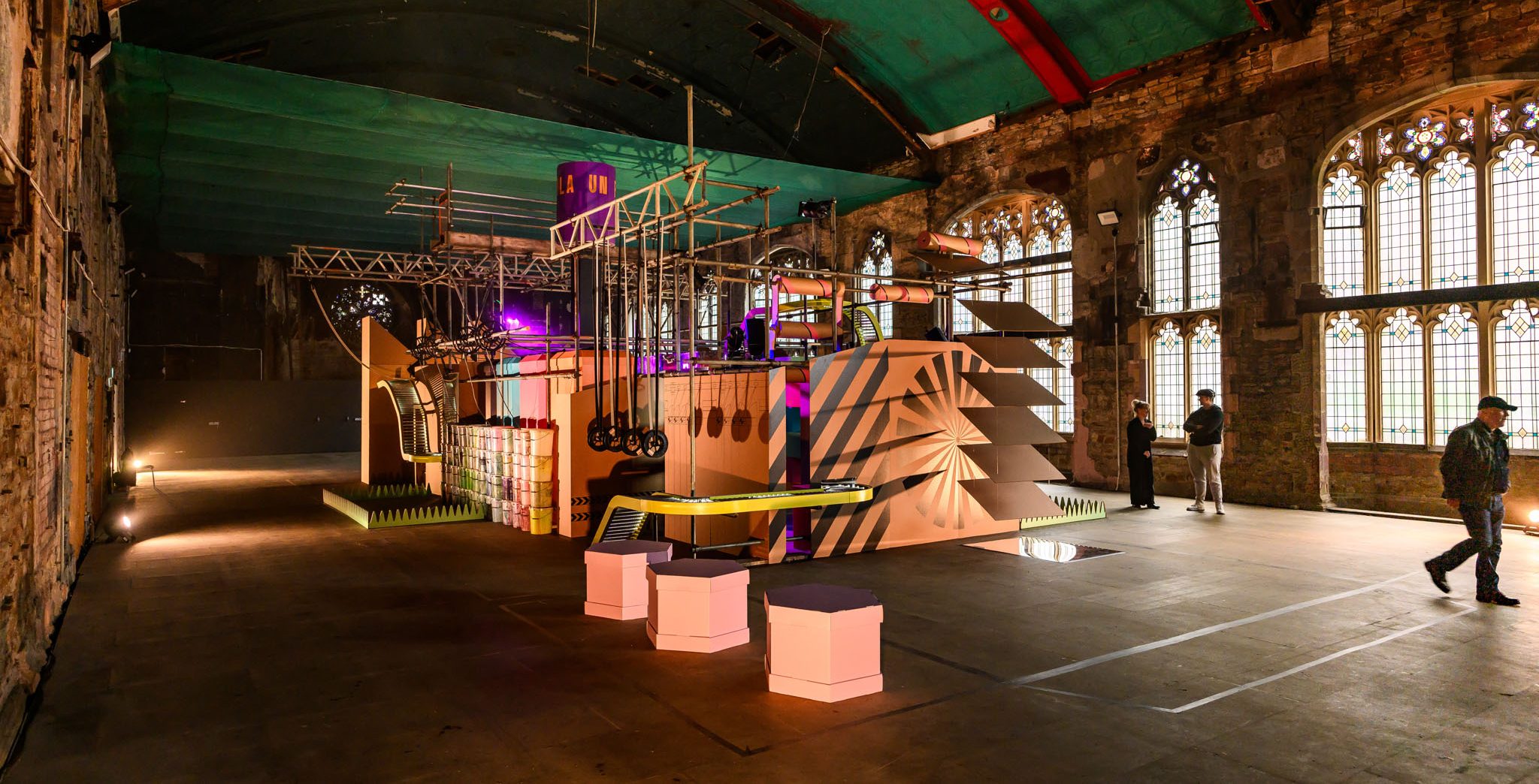 A brightly lit cardboard art installation of ropes and bars sits in a large industrial hall with arched windows and exposed brick, as visitors observe.