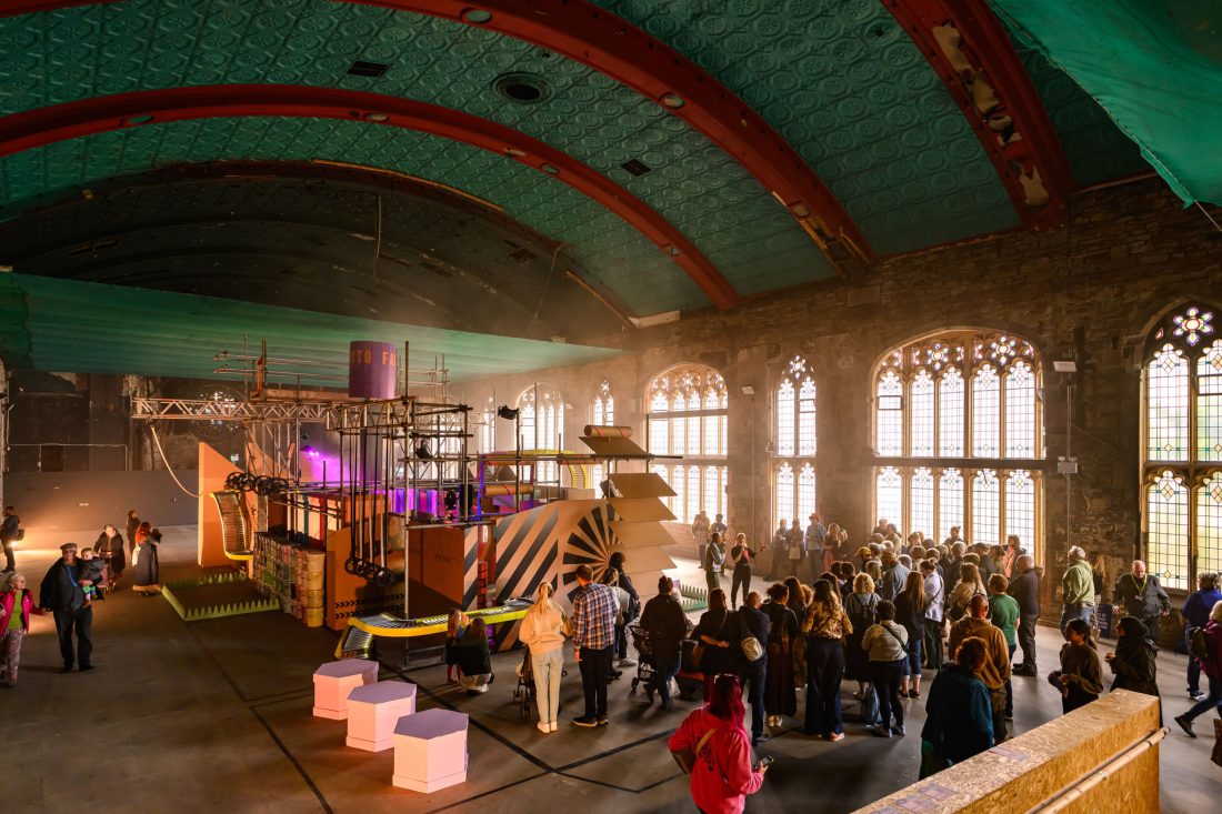 A large crowd gathers inside a grand hall with tall arched windows, ornate ceiling, and a large cardboard art installation at the centre. Sunlight streams in, illuminating the people and the artwork.