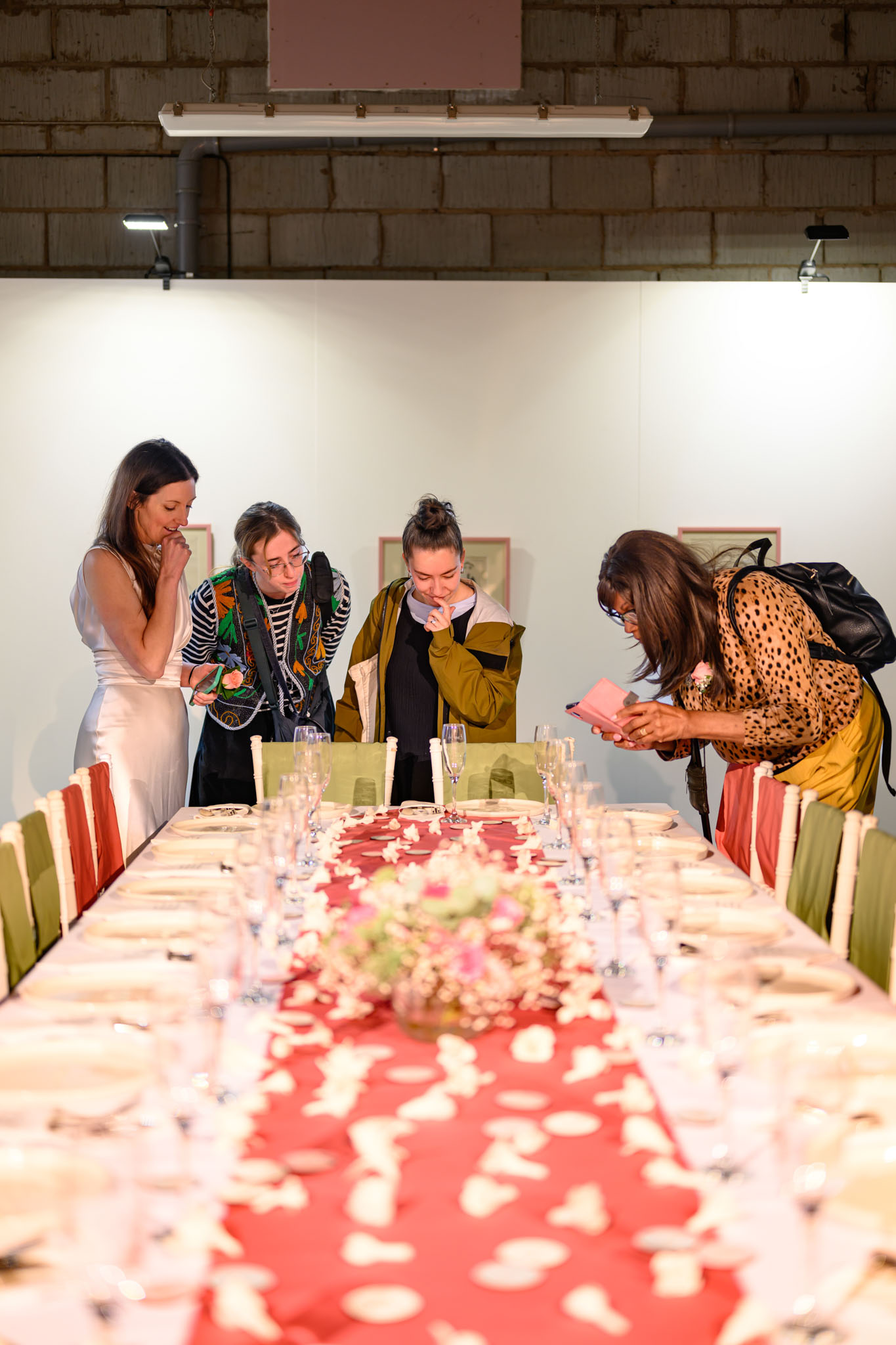 Four women stand at the end of a long, formal dining table with a red and white table runner, looking closely and discussing something on the table, which is set with plates, glasses, and a floral centrepiece.
