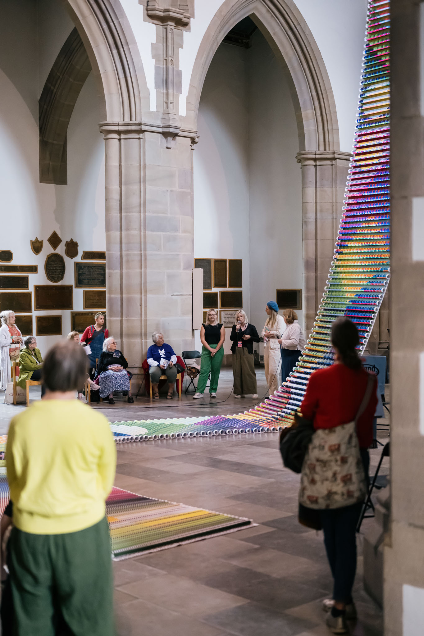 People observe and discuss a tall, colourful, rainbow art installation made of small objects inside a large stone-arched building, possibly a church or gallery. Several people are seated or standing around the artwork.