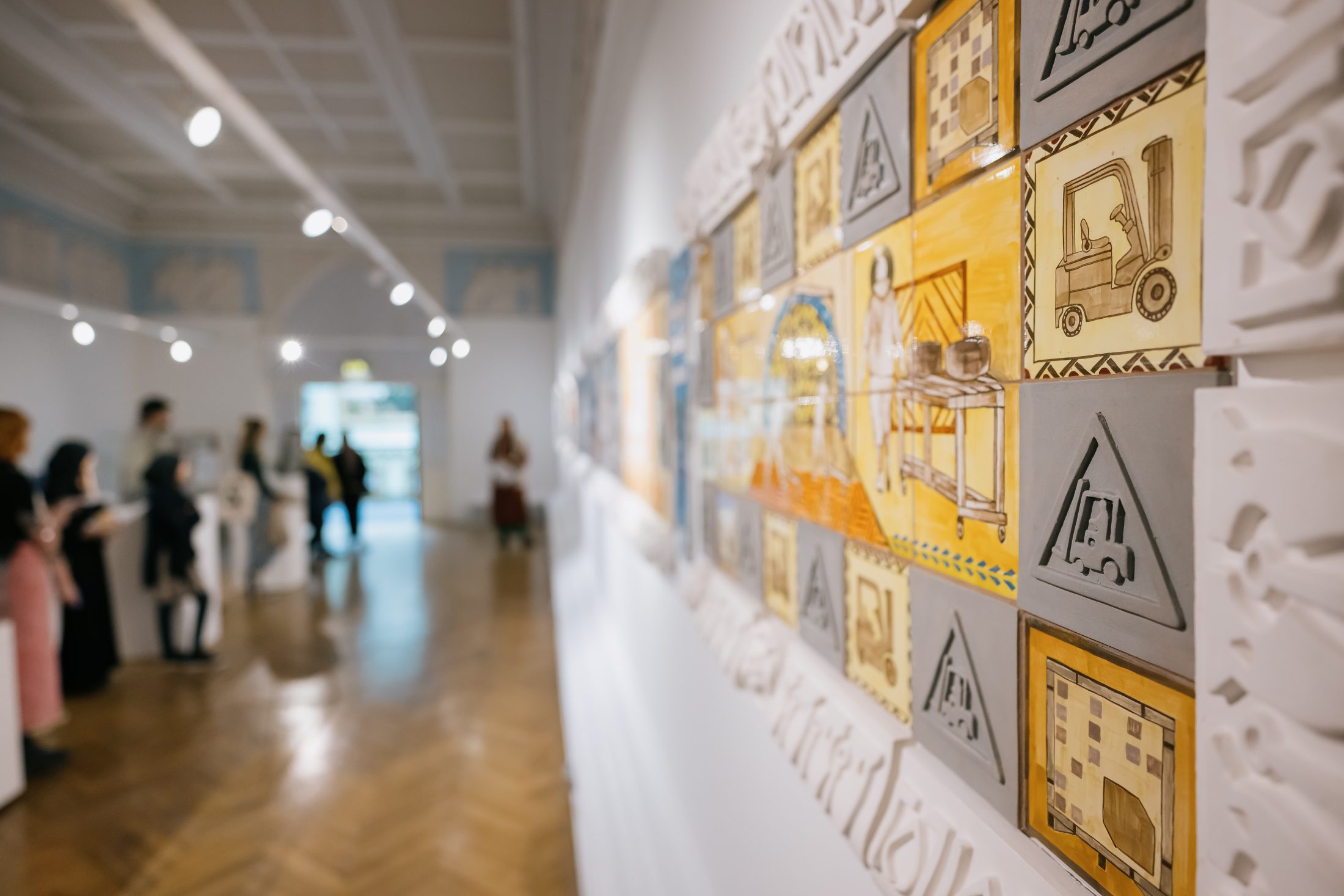 A close-up of colourful, industrial-themed artwork on a gallery wall, with visitors viewing other exhibits in the blurred background of a well-lit art museum.