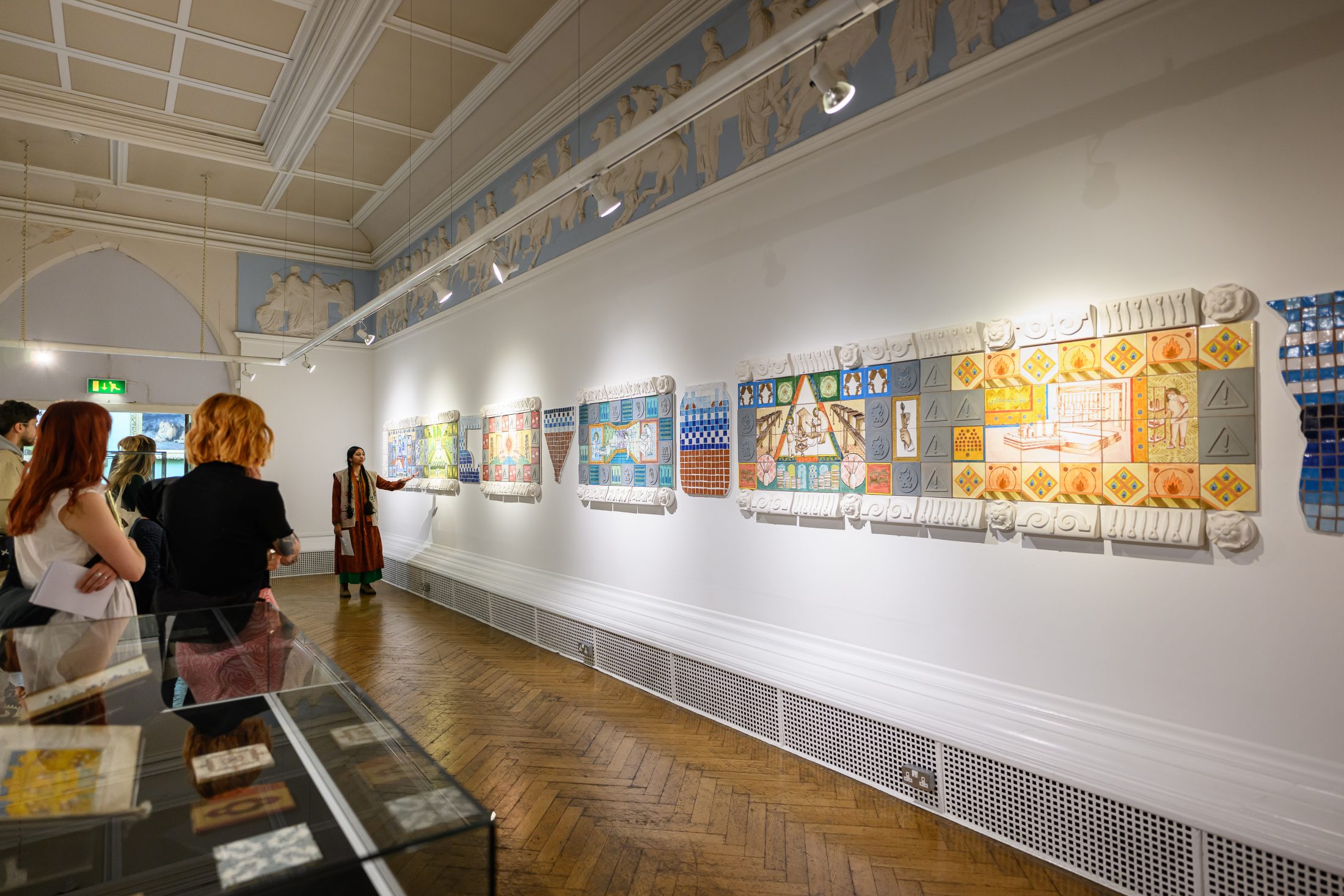 Visitors observe colourful, tile artworks displayed on a white gallery wall in a well-lit exhibition space with wooden parquet floors and classical decorative mouldings on the ceiling.