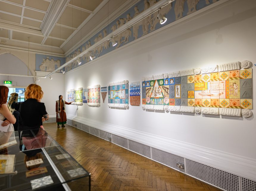 Visitors observe colourful, tile artworks displayed on a white gallery wall in a well-lit exhibition space with wooden parquet floors and classical decorative mouldings on the ceiling.