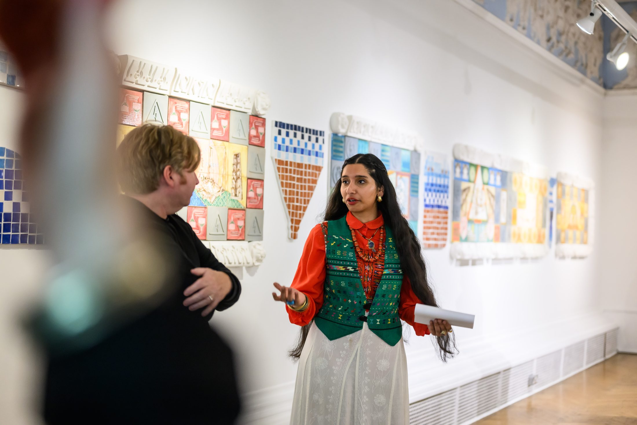 Two people stand and talk in an art gallery, with colourful tiled artworks displayed on the white walls behind them. One person gestures while holding papers, and the other listens with arms slightly crossed.
