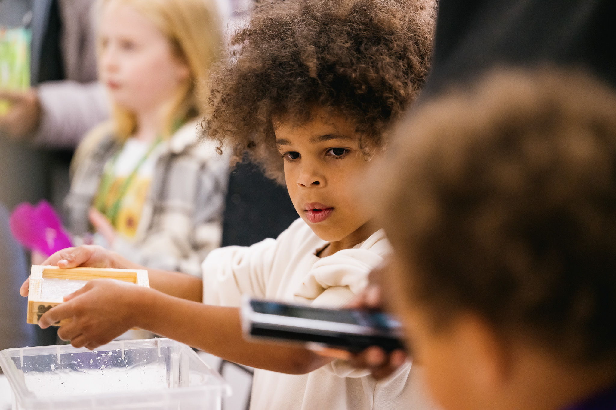 A young child with curly hair holds a box while taking part in an activity. Other children and adults are nearby, with one child blurred in the foreground. The setting appears to be a classroom or group event.