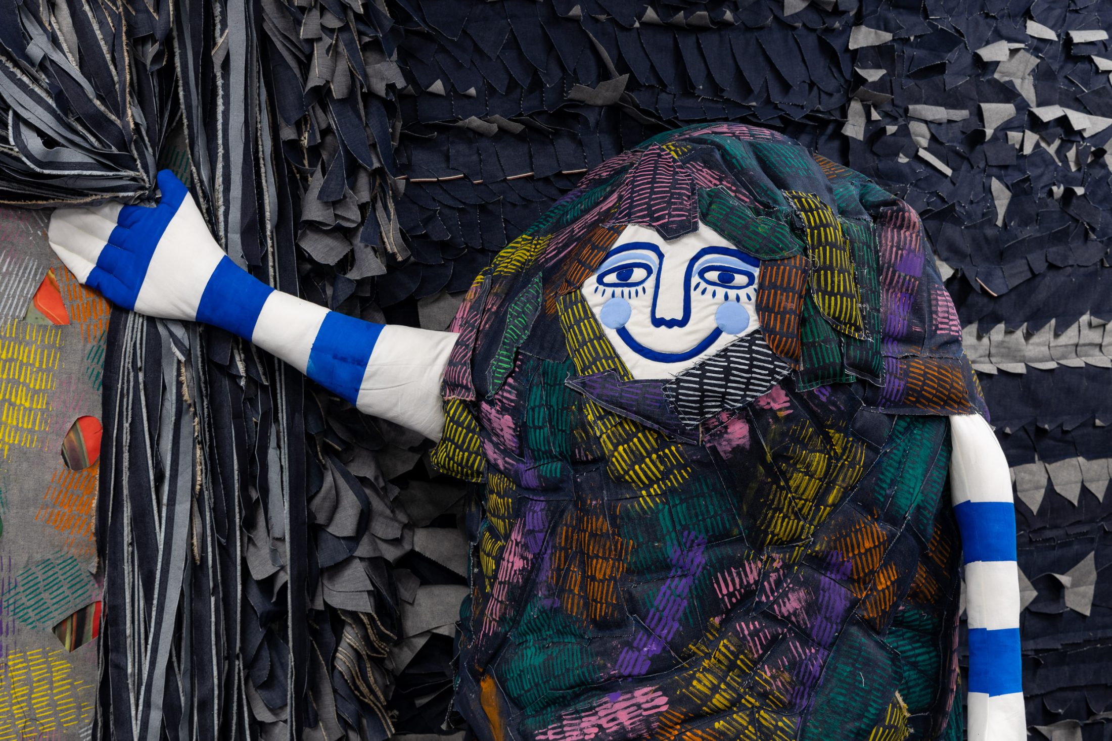 A colourful, fabric figure with a blue and white striped arm, a smiling face, and a multicoloured patchwork body holds a curtain against a textured, dark, layered background.