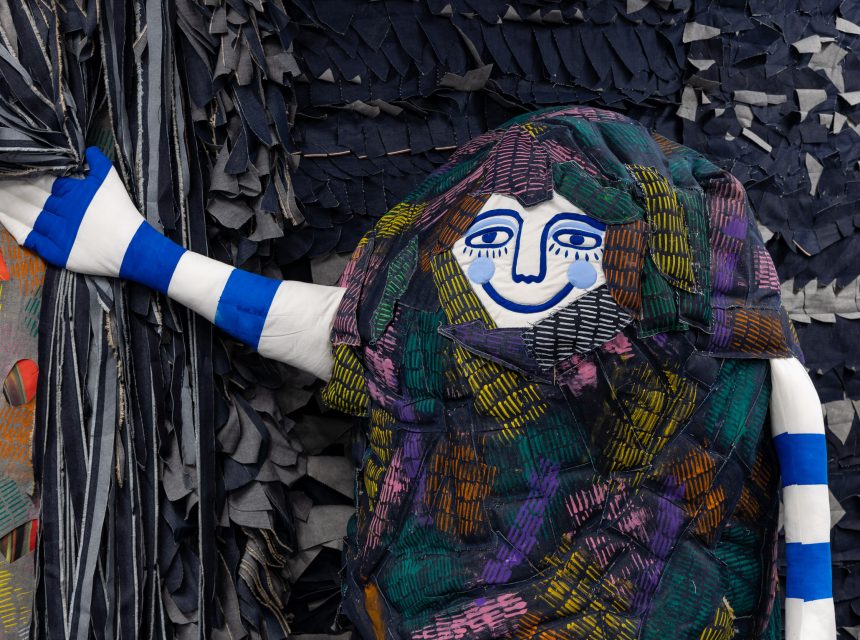 A colourful, fabric figure with a blue and white striped arm, a smiling face, and a multicoloured patchwork body holds a curtain against a textured, dark, layered background.