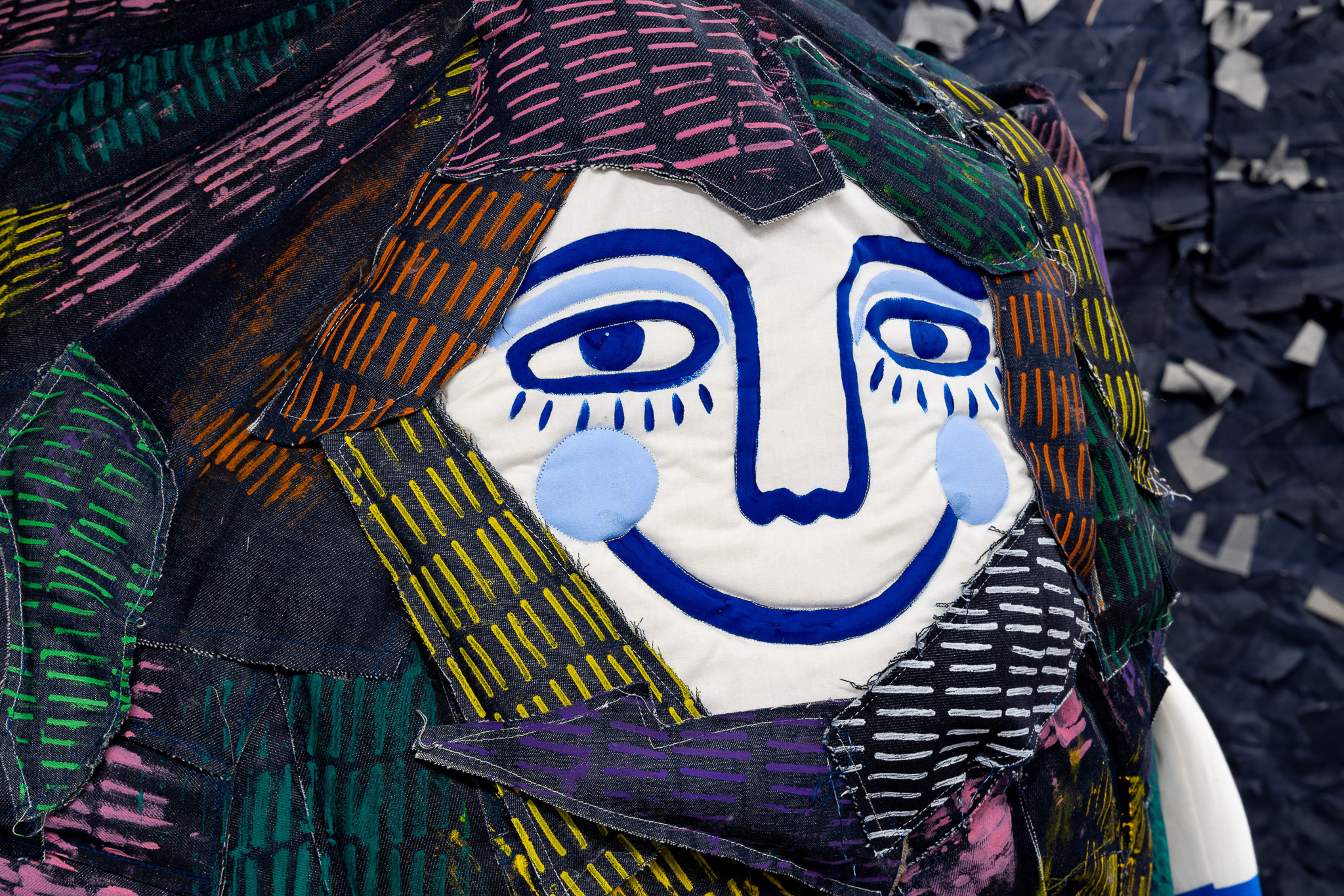A close-up of a colourful fabric sculpture featuring a smiling face with bold blue features, blue cheeks, and patterned patches of fabric in various colours forming hair around the face.