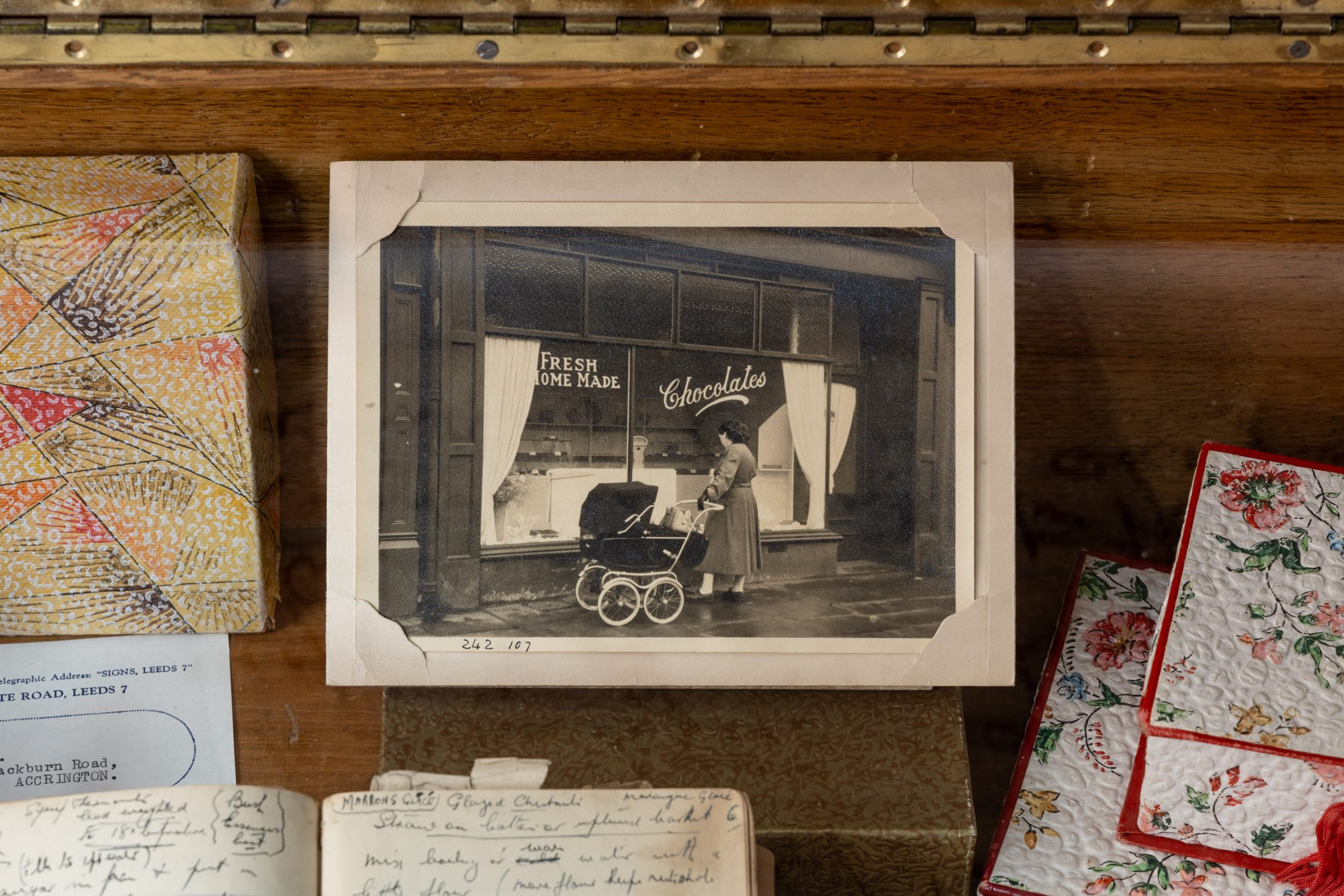 A black-and-white photo shows a woman with a pram outside a shop window displaying the sign Fresh Home-made Chocolates. The photo is placed among vintage letters, a notebook, and embroidered cloths.