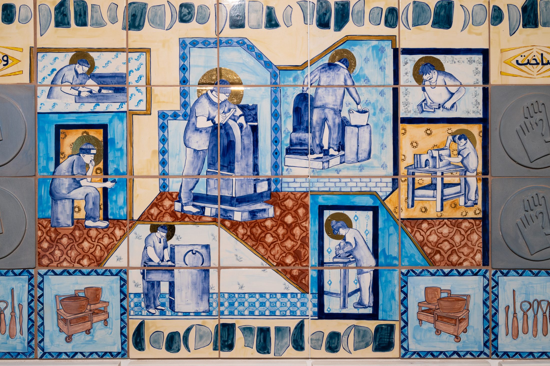 Colourful ceramic tile mural illustrating people engaged in pottery-making and sculpture, depicted in blue and gold tones, with patterned borders and small images of pottery and tools surrounding the scenes.