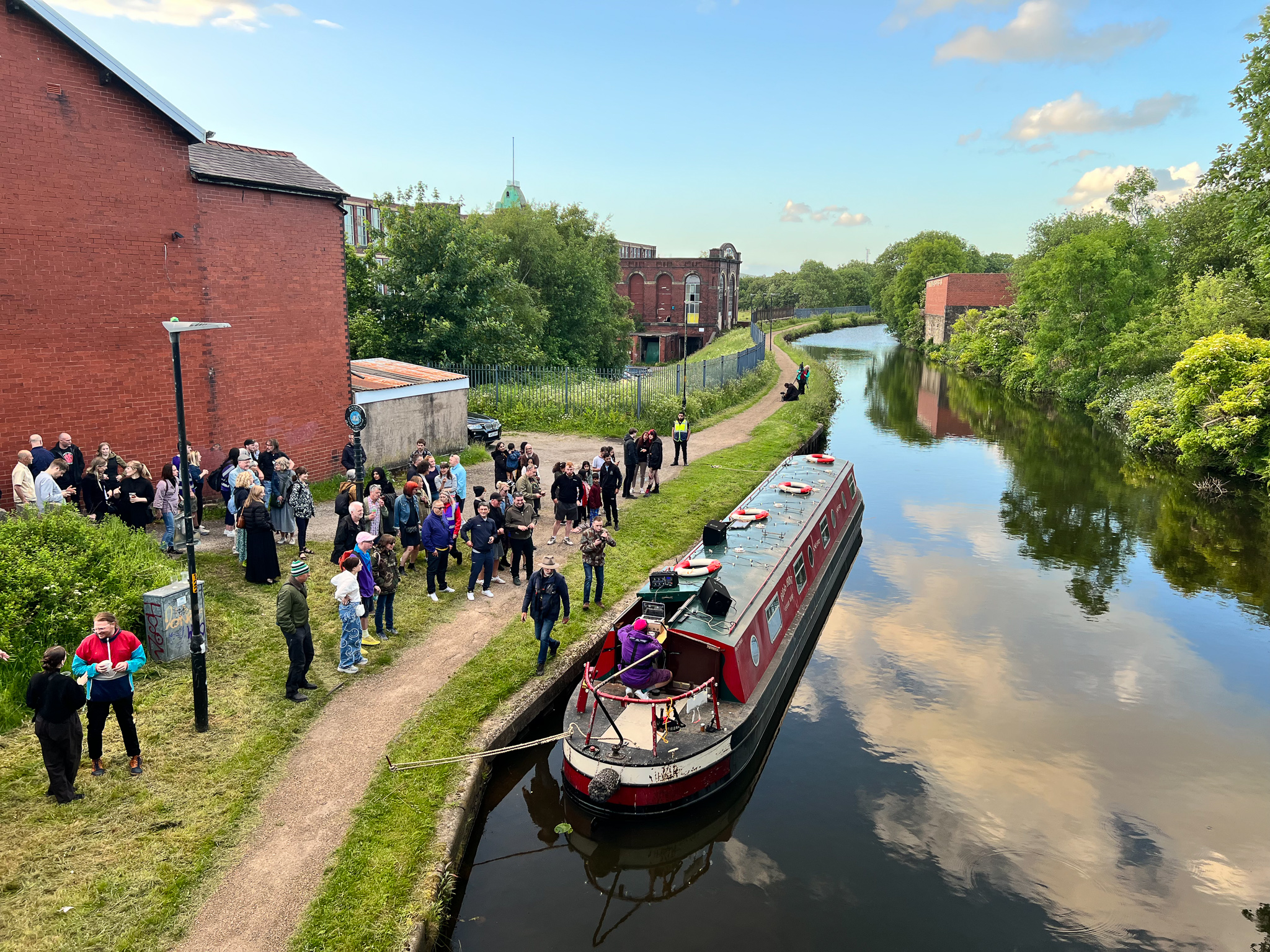 Red canal boat moored on a calm river under blue skies with fluffy clouds, as a group of people stand on a grassy path by brick buildings enjoying the scene.