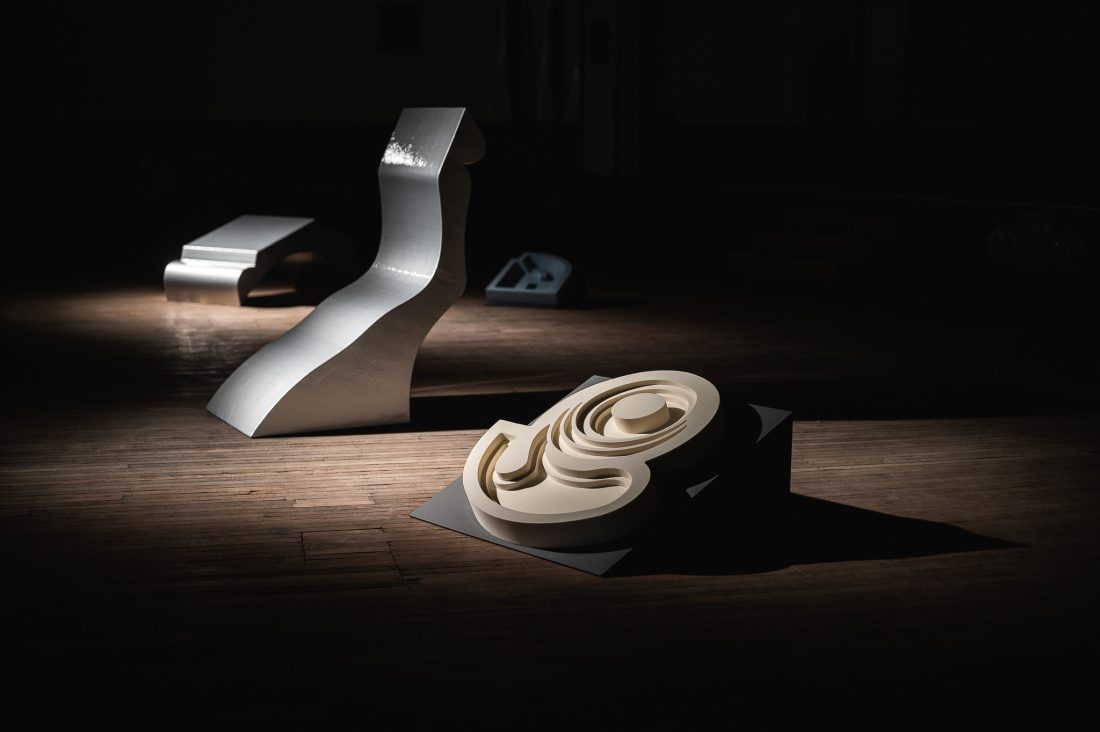 Three abstract sculptures in a dimly lit room: one silver and curved, one geometric and metallic, and one cream-coloured with layered, swirling shapes, all set on a wooden floor with dramatic spotlighting.