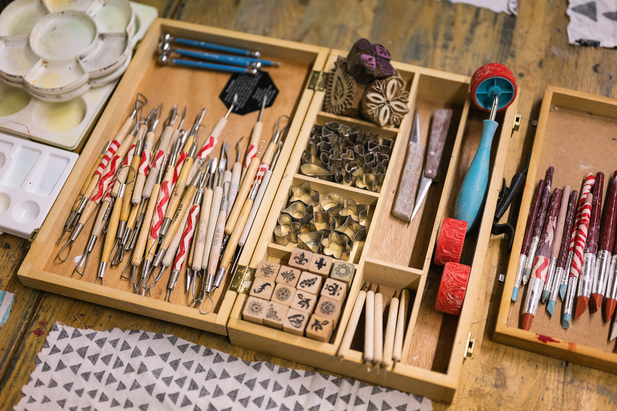 A wooden box filled with pottery and clay tools, patterned stamps, carving tools, rollers, brushes, and metal cutters, organised neatly on a wooden table with fabric swatches nearby.