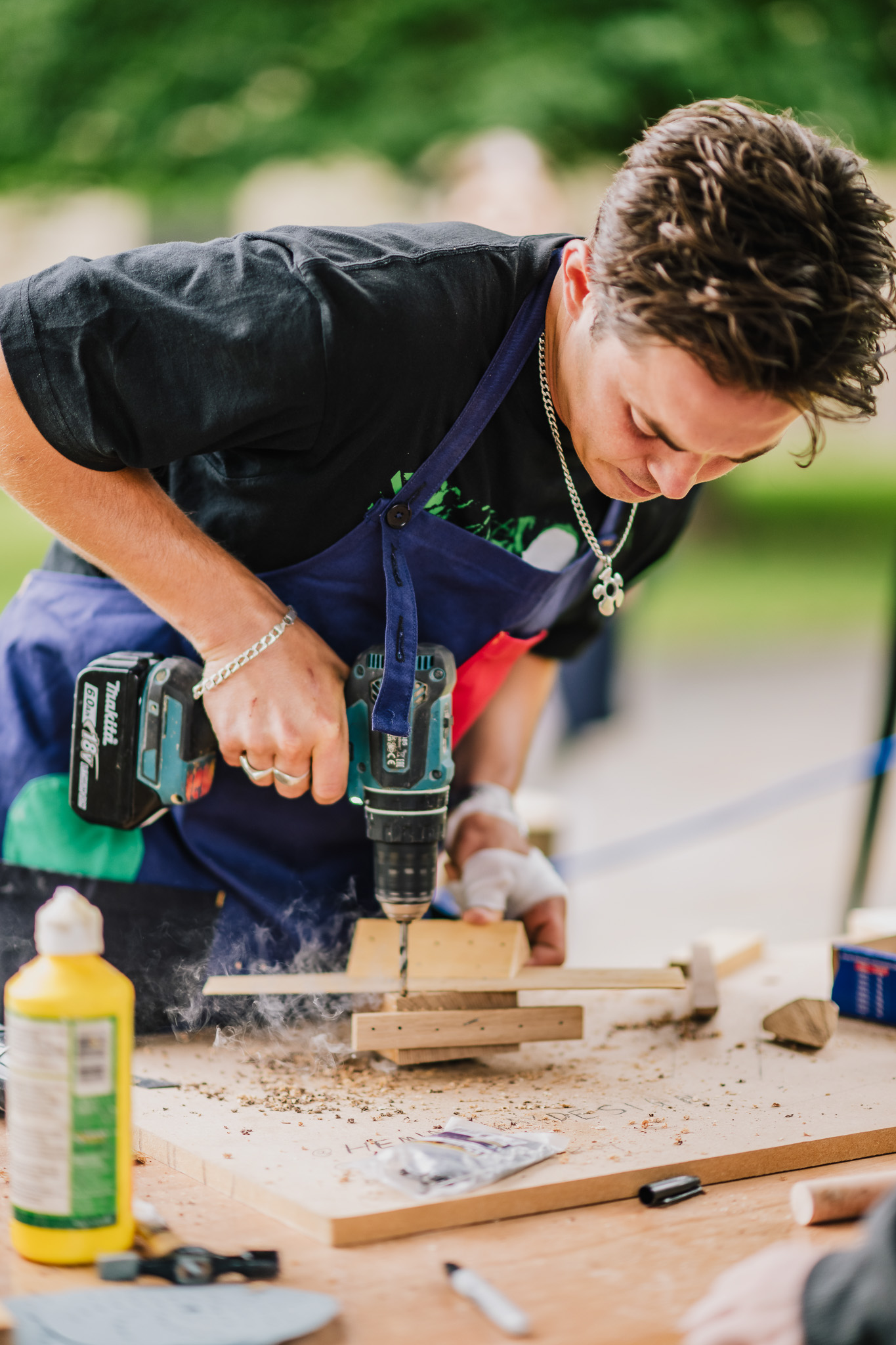 A man in a blue apron uses a power drill on wood, surrounded by tools and sawdust, in an outdoor setting.