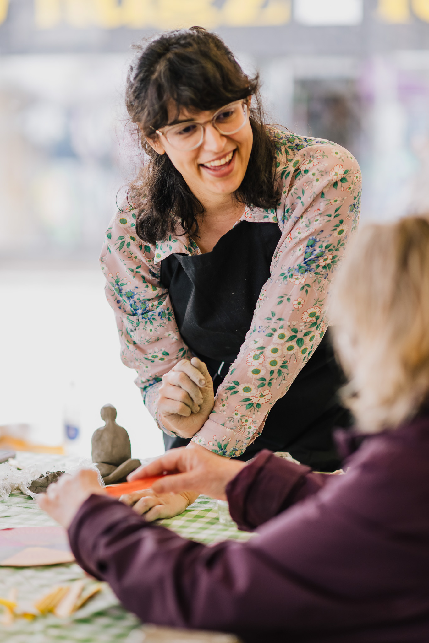 Smiling woman in floral shirt and apron teaches pottery to a seated person, with clay figures on a checkered table.
