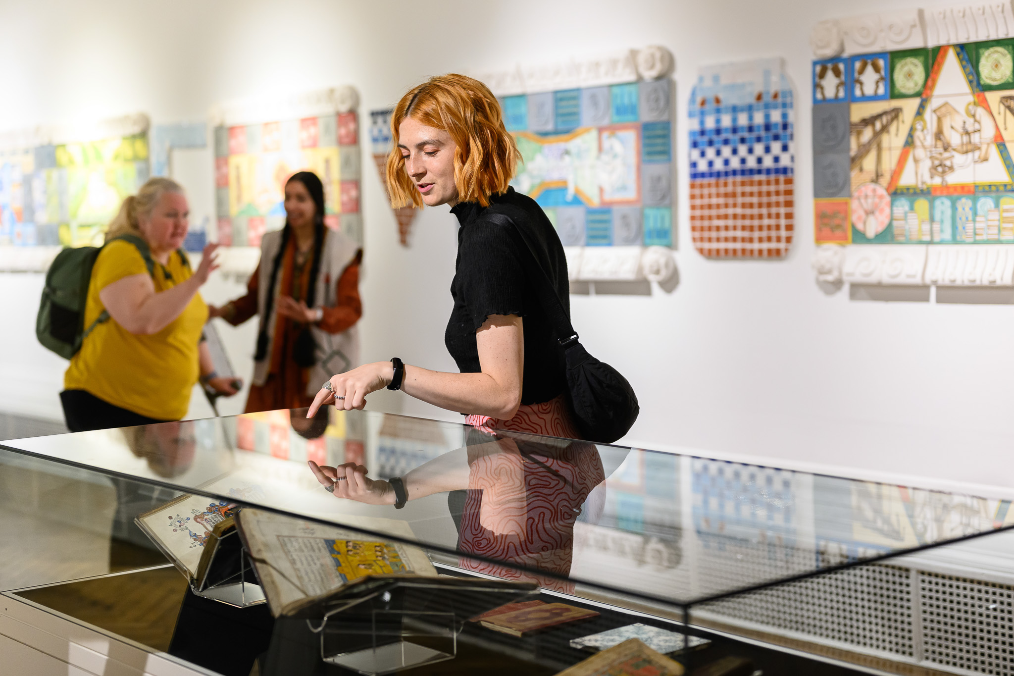 A woman with red hair looks at objects in a glass display case at an art gallery, with colourful artworks hung on the white walls behind her. Two other women are visible in the background.