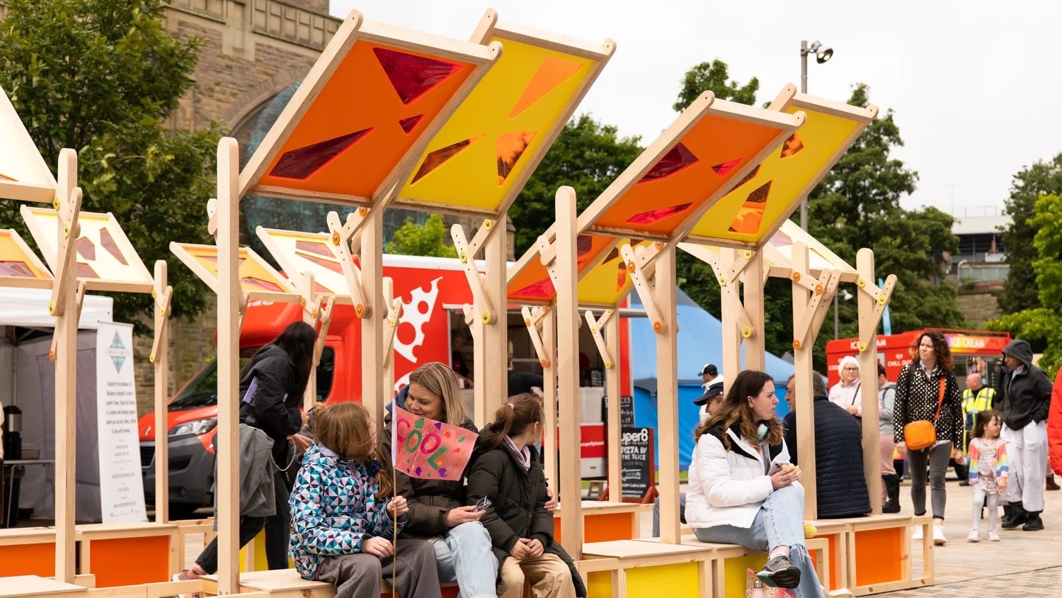 People sit and chat under colourful, geometric sunshades on wooden benches at an outdoor event, with food vans and other attendees visible in the background. Trees and buildings frame the lively scene.