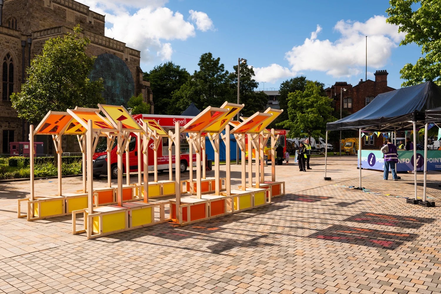Colourful wooden market stalls with orange and yellow accents stand empty on a paved outdoor area. Nearby, people gather under tents labelled “INFO POINT,” with buildings and trees in the background under a blue sky.