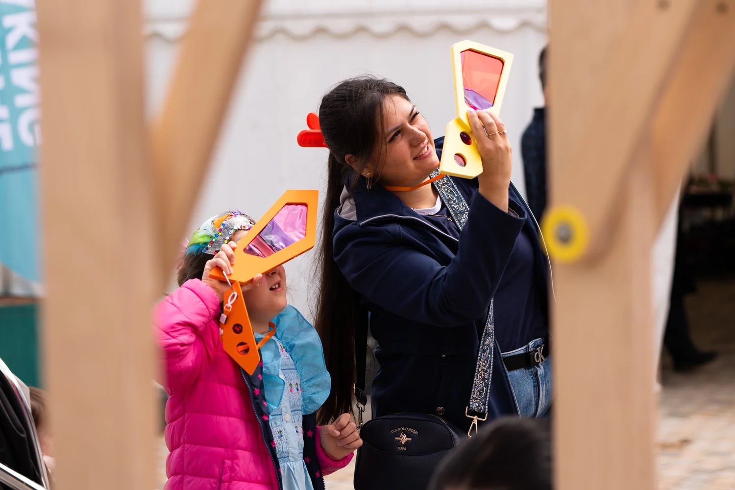 A woman and a child smile whilst holding up colourful kaleidoscope viewers, looking through them at an outdoor event. The child wears a pink jacket and headband; the woman wears a navy jacket and jeans.