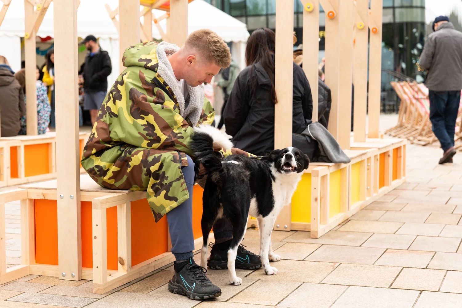 A man in a camouflage jacket sits on a bench and strokes a black and white dog, who looks back with a happy expression. People walk and gather in the background at an outdoor event.