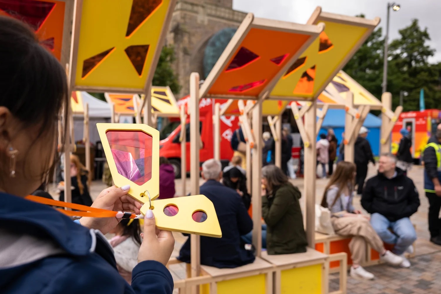 A person holds a colourful, geometric, translucent panel at an outdoor event, where people sit and gather under matching vibrant, modern wooden structures. Bright orange and yellow panels provide shade.