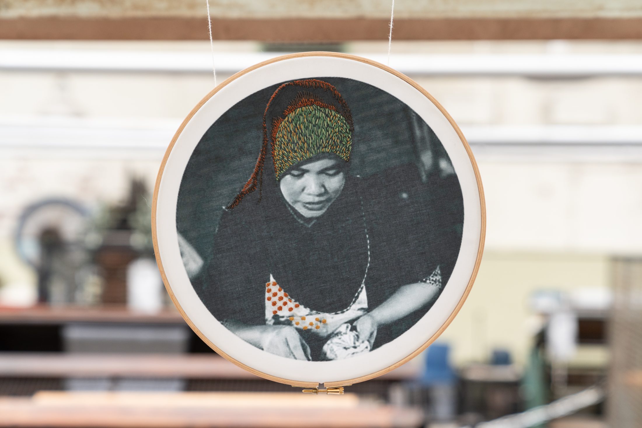 A circular embroidery hoop displays a black-and-white image of a woman wearing a headscarf and patterned clothing, with colourful thread accentuating her headscarf. She appears to be focused on an activity with her hands.