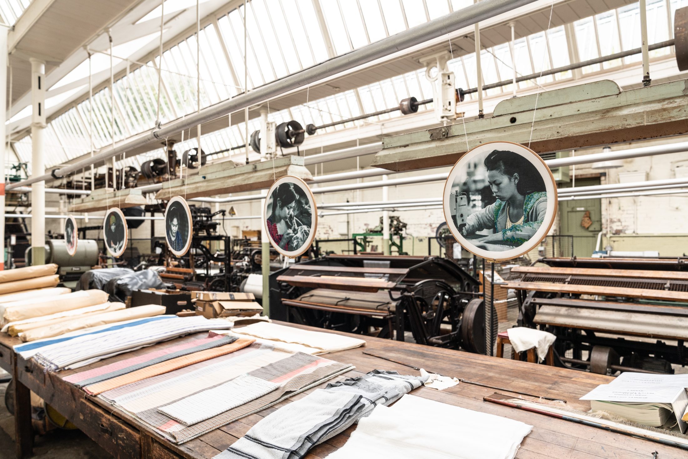 A textile factory interior with rolls of fabric on a wooden table and vintage machinery in the background. Round signs with black-and-white portraits of workers hang above the machines. Natural light streams from a slanted glass roof.