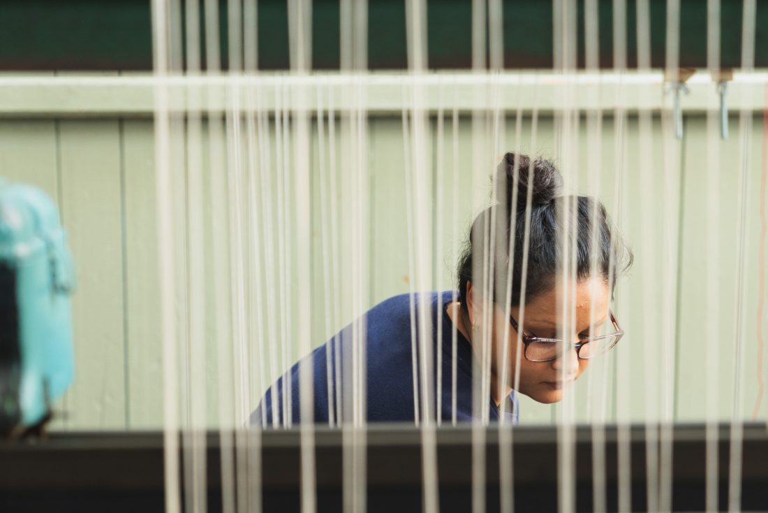 A person with glasses and dark hair in a bun is focused on their work, seen through vertical threads on a loom. The background is a light green wall.