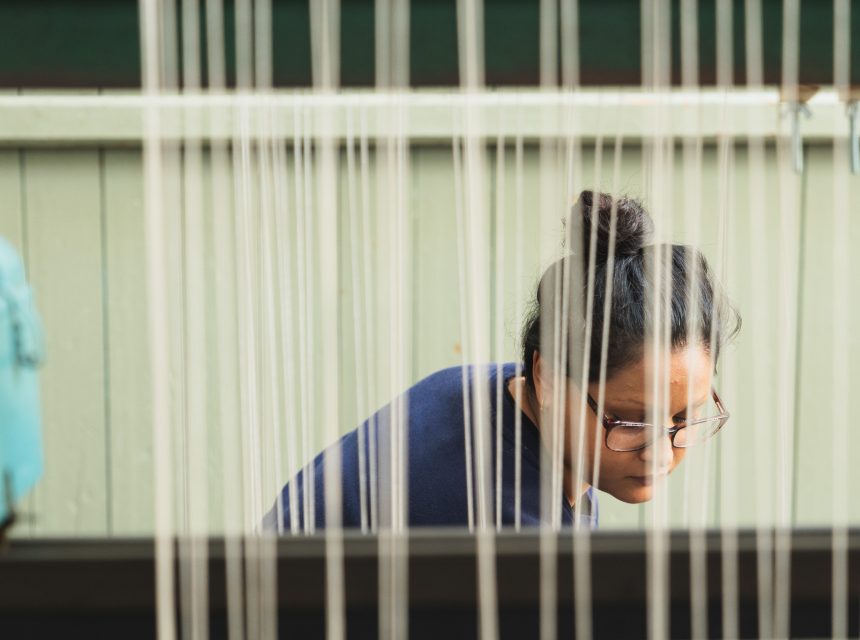 A person with glasses and dark hair in a bun is focused on their work, seen through vertical threads on a loom. The background is a light green wall.