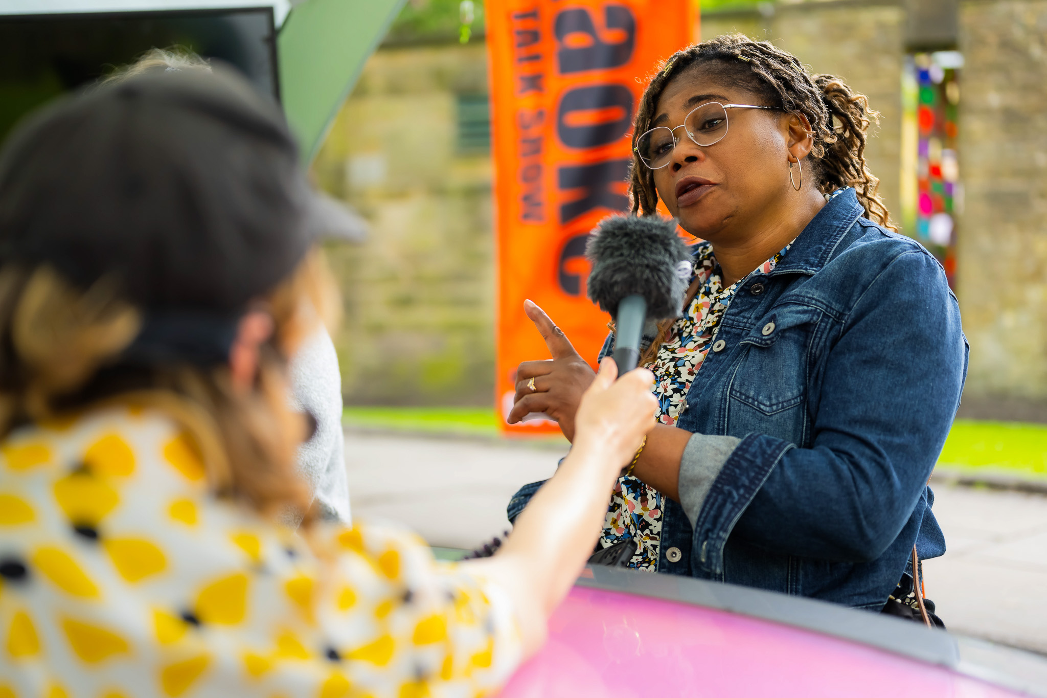 A woman in a denim jacket speaks passionately whilst being interviewed outdoors by a person holding a microphone. An orange banner and a stone building are visible in the background.